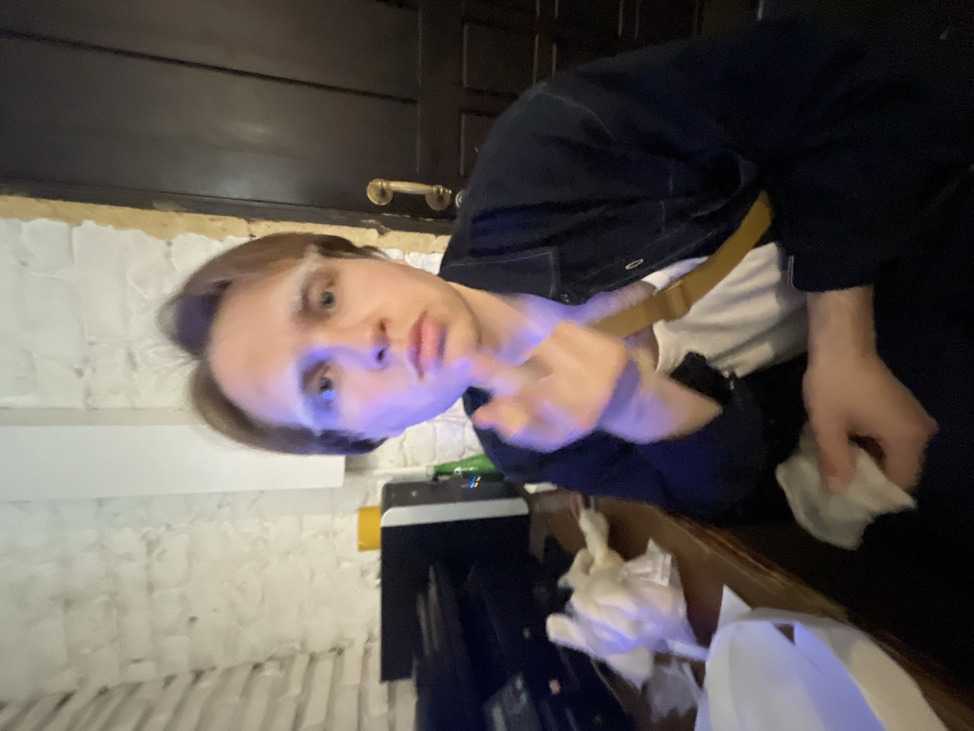 A young man is holding a piece of paper in his right hand, while his left hand is raised. He is wearing a dark blue jacket over a white t-shirt. There is a tan strap visible across his chest, possibly from a bag. The background includes a white brick wall, a dark door with a gold handle, a computer monitor, and a pile of tissues or paper napkins on a wooden surface. Based on the stucco-like texture of the wall and the interior design, the location may be a bar or restaurant, but the city cannot be confidently determined from this image alone. More context or specific landmarks would be needed to identify the location as Moscow, Russia.