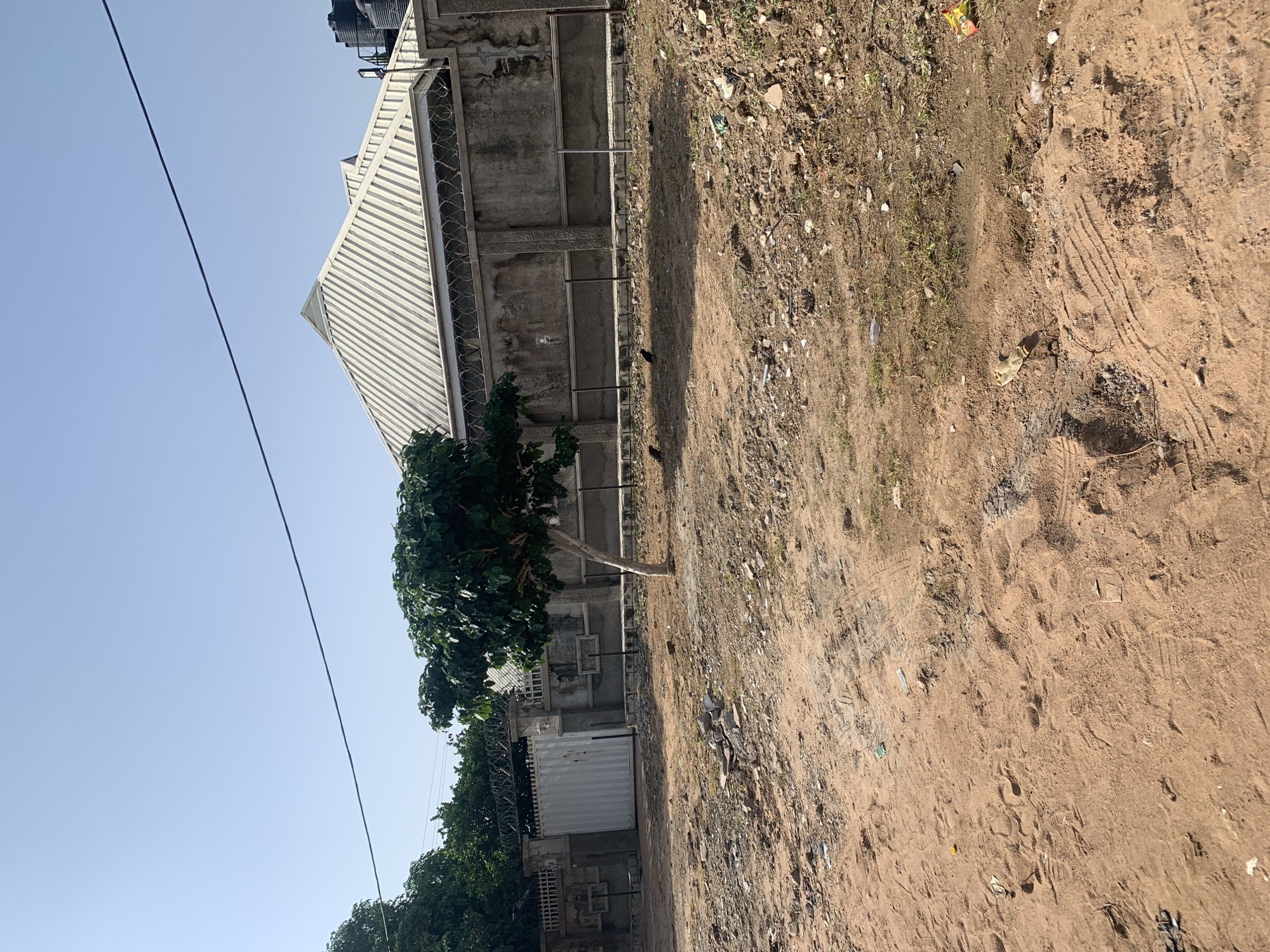An outdoor, daytime, wide shot captures a residential property with a concrete wall topped with barbed wire fencing. The wall surrounds a building with a silver, ridged roof. A large tree with dense green foliage stands in front of the wall, to the left of center. The ground in the foreground is a mix of dirt and scattered debris. The sky is clear and blue.