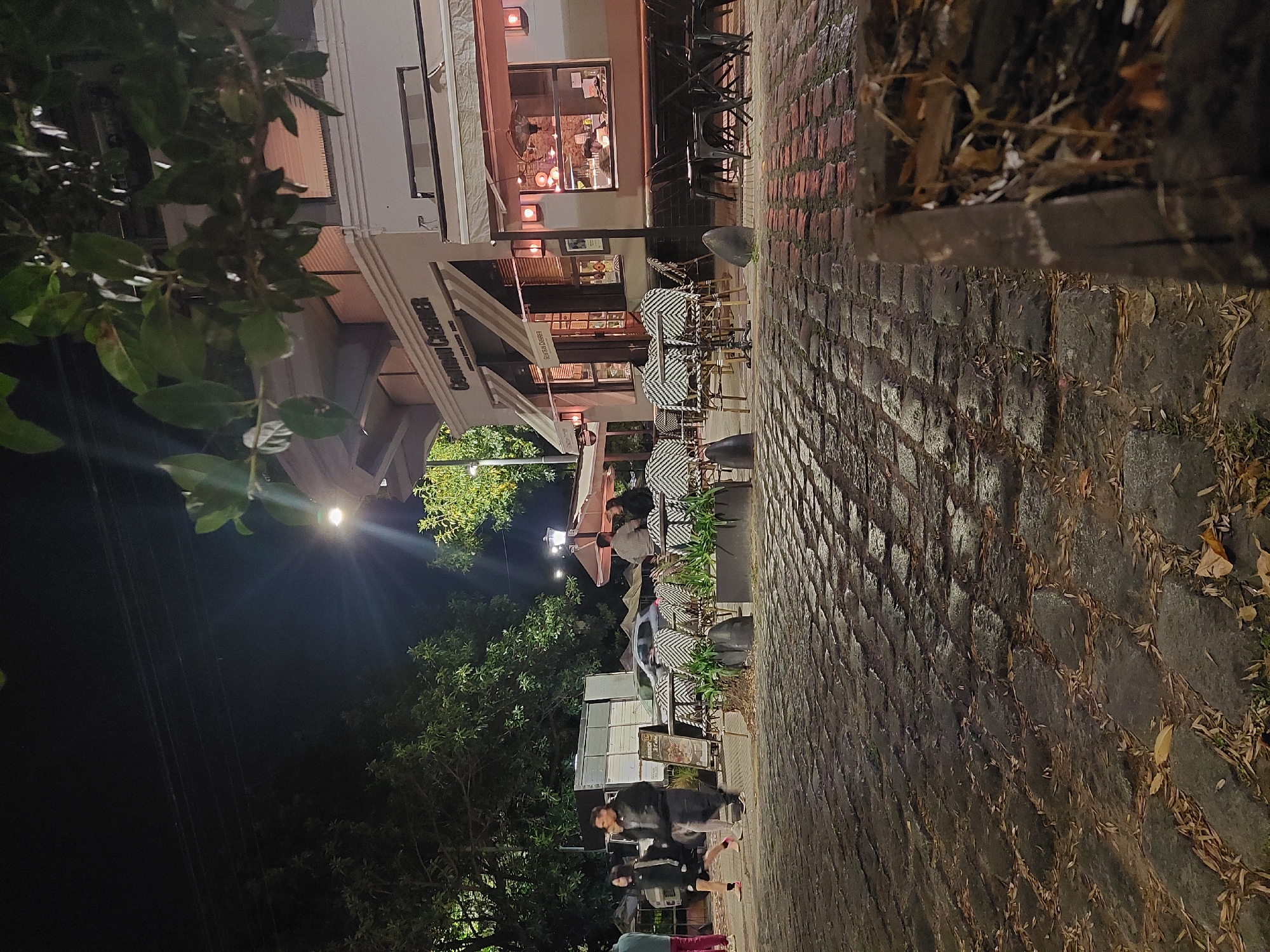 At night in Buenos Aires, Argentina, people walk on a cobblestone sidewalk adjacent to a building with a cafe or restaurant. Tables and chairs are set up outside, and some people are seated. The building has awnings and lights, and trees are visible in the background. A street light illuminates the scene.