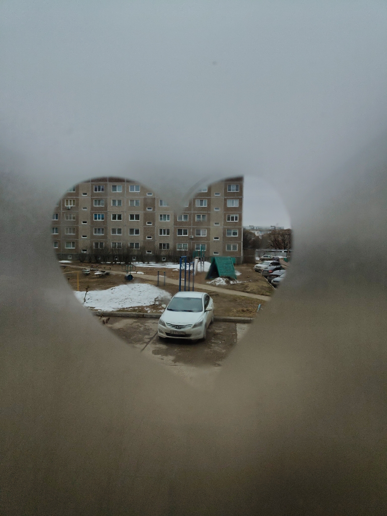 The image shows a view of a building and a parking lot seen through a heart-shaped opening. The building is a multi-story apartment complex with rows of windows. In the parking lot, a white car is parked. Patches of snow are visible on the ground. In the background, additional buildings and vehicles are visible. The heart-shaped opening is somewhat blurred, suggesting it is part of a foreground object that is close to the camera. The sky appears overcast. The location is Unknown, Unknown.