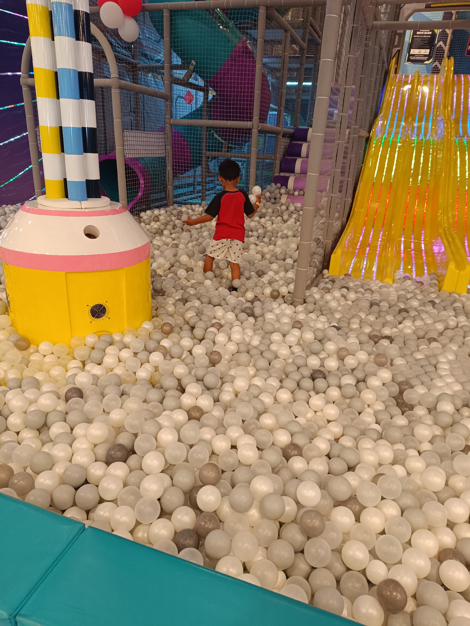 This is a medium-angle, eye-level, full shot of a young boy playing in a large ball pit. The boy, wearing a red shirt and patterned shorts, is facing away from the camera and is holding a white ball in his right hand. He is standing in a ball pit filled with hundreds of white and gray balls. To the left of the boy is a large, yellow, cylindrical structure with a white and pink top. In the background, there is a colorful, winding yellow slide with multicolored lights. The environment appears to be an indoor playground. The image was taken in Sorong, Indonesia.