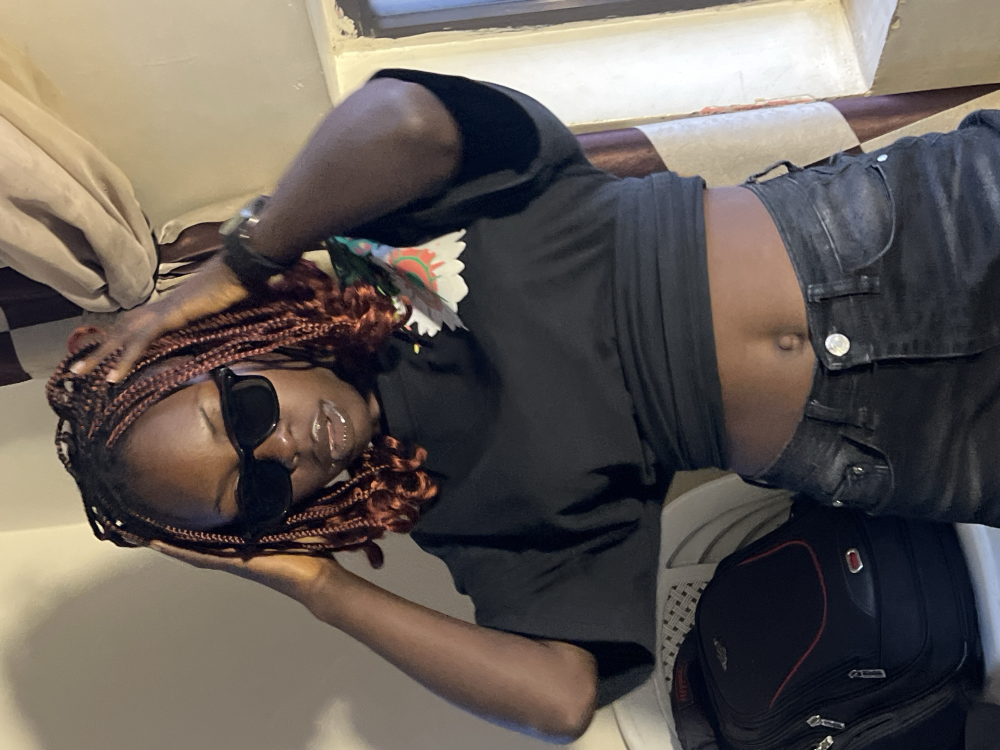 A young Black woman with dark skin and braided auburn hair is standing indoors, posing with her hands on her head. She is wearing a black crop top and dark wash jeans, sunglasses, and lip gloss. A black backpack is visible on the left side of the frame. The background includes a wall, a curtain, and a window. The lighting suggests an indoor setting with artificial light and some natural light coming from the window. The woman appears to be in a relaxed and confident pose. There is no specific information in the image to suggest the location is in Jos, Nigeria.