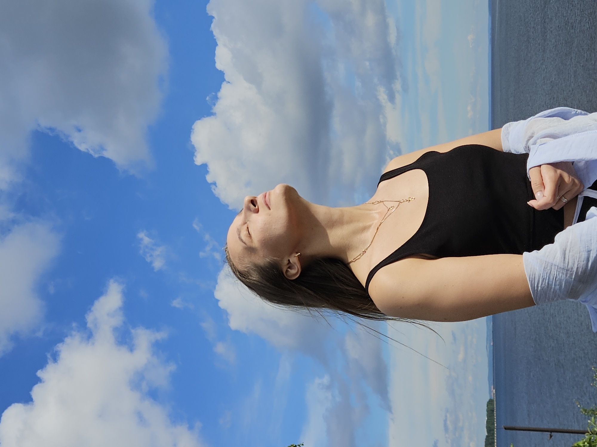 A young woman with long brown hair, wearing a black tank top and a light blue open shirt, stands outdoors with her eyes closed and face tilted towards the sky. She is adorned with a gold necklace and a ring on her right hand. The background features a vast blue sky filled with white and gray clouds, a calm body of water, and a distant tree line. The scene appears to be a scenic overlook, possibly near the Kama River within the city of Chaikovsky, Russia, given the natural landscape elements.
