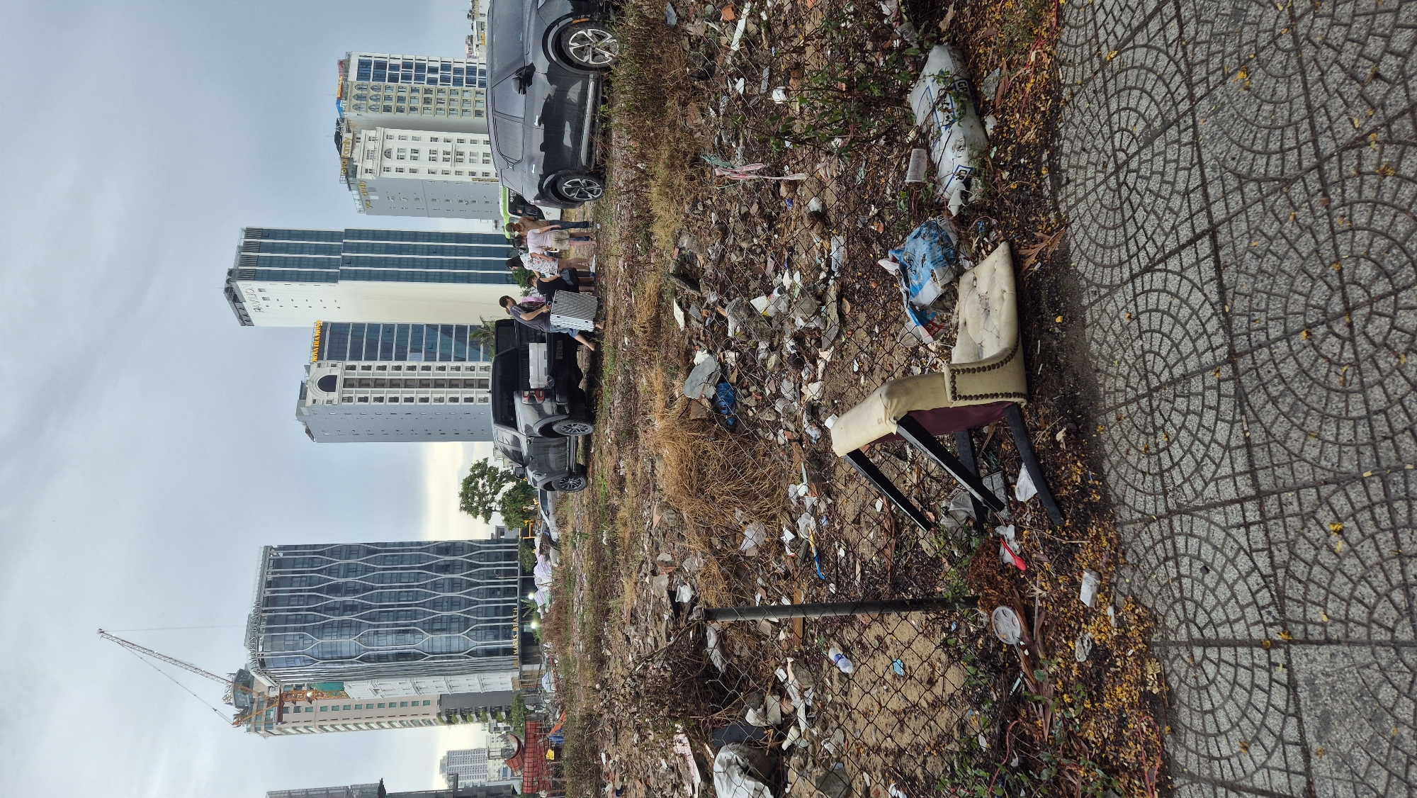 The image depicts a scene of urban decay and neglect in Đà Nẵng, Vietnam. The foreground is dominated by a cobblestone pavement showing signs of wear and tear, with scattered yellow leaves and debris. Beyond the pavement, a chain-link fence encloses a desolate, trash-strewn lot. The ground is littered with plastic bottles, bags, construction debris, dried vegetation, and a broken chair with a cream-colored seat and a dark red backrest.
In the middle ground, a black pickup truck is parked, its tailgate down, with several people gathered around it. A woman is seen carrying a suitcase, and others appear to be involved in loading or unloading items. Next to the pickup truck is a black SUV.
The background features a cluster of modern high-rise buildings under a cloudy sky, indicative of an urban environment. One building on the left is under construction, with a prominent crane. The contrast between the sleek, contemporary architecture and the squalid, neglected foreground is stark. The overall impression is one of a city undergoing development, but with pockets of blight and disarray.