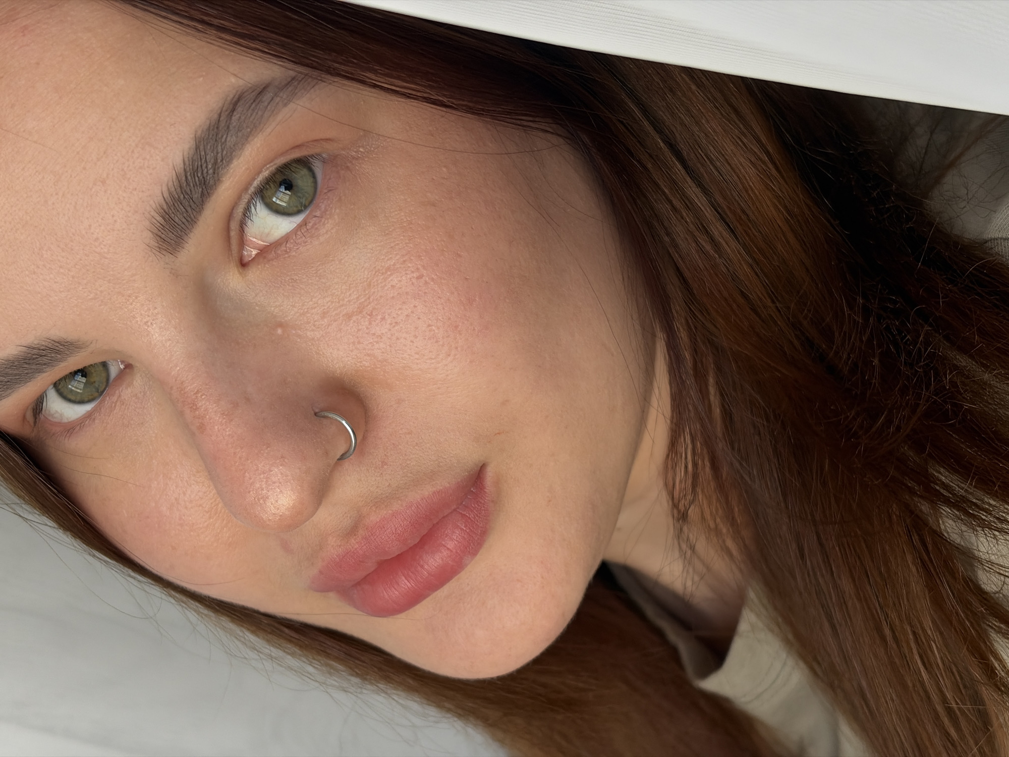 A close-up shot of a woman's face, angled towards the camera. She has fair skin, light brown hair, and green eyes. A silver hoop is visible in her left nostril. The setting appears to be indoors with indirect lighting. The city and specific location are unknown.