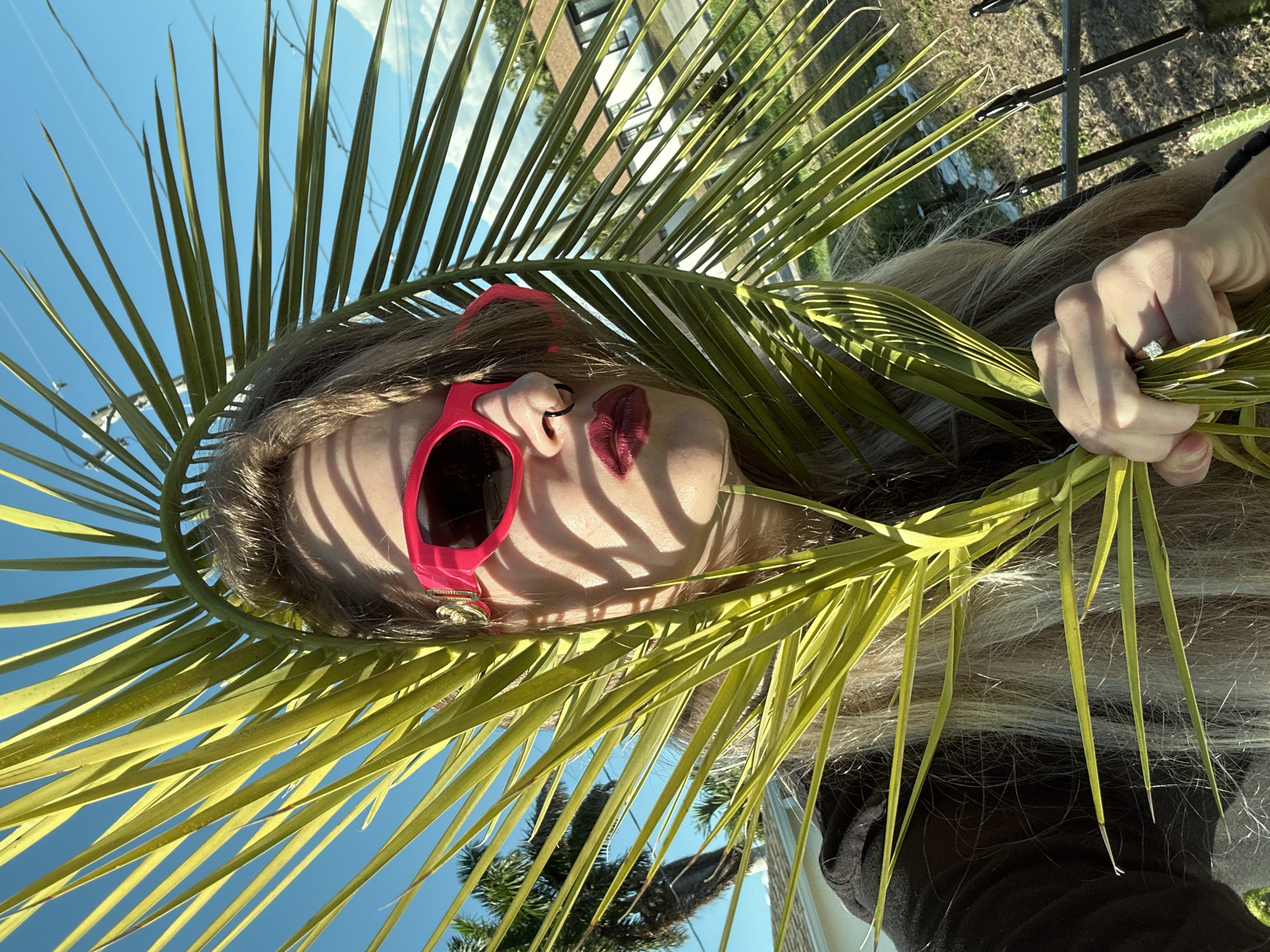 A woman is holding a palm frond in front of her face. She is wearing red sunglasses and has lipstick on. Her hair is blond and she is wearing a ring on her finger. The sky is blue and the building is a two-story structure. The image was likely taken in the city of Cape Coral, United States of America.