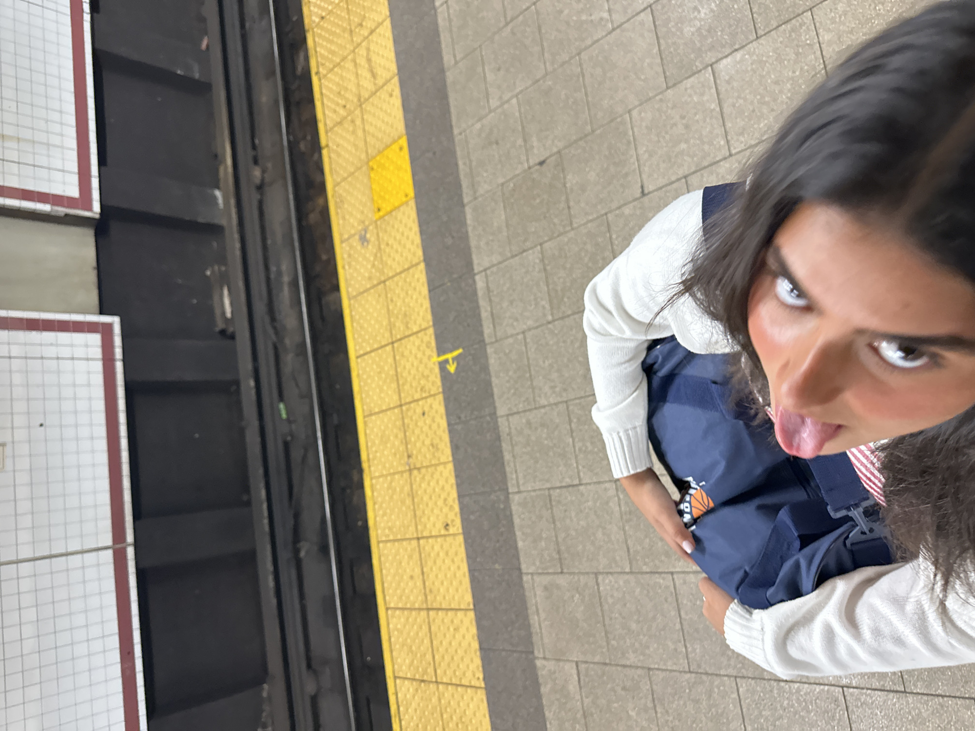 A high-angle shot captures a young person posing on a subway platform. The individual has long, dark hair and is looking directly up at the camera with an intense, wide-eyed gaze, playfully sticking their tongue out. They are wearing a white long-sleeved knit top and dark blue overalls or a similar garment.
A blue bag, possibly a backpack or duffel, is attached to the front of the blue garment, featuring a partially visible circular orange logo with a basketball design. The person's fingernails appear to have a light pink or nude polish.
The setting is a subway station platform, characterized by light-colored square floor tiles. To the left of the person, a bright yellow tactile warning strip runs along the edge of the platform. Beyond this strip are the dark subway tracks and a gap. A white tiled wall with distinctive red border lines, common in New York City subway stations, is visible in the background to the far left.
A small, faint yellow arrow symbol, pointing downwards with a short horizontal line, is visible on the yellow tactile strip. The time of day is not discernible due to the indoor setting with artificial lighting. The overall mood conveyed is playful and energetic. Given the visual cues, the location is strongly indicated as a New York City subway station.