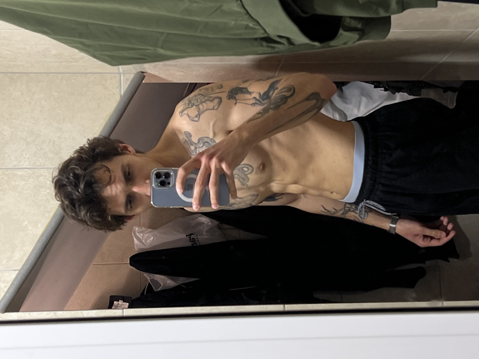 A male individual with dark, curly hair and a lean, muscular physique is depicted taking a mirror selfie indoors. He is shirtless, displaying multiple tattoos across his chest, shoulders, and arms, and wears black athletic shorts with a light blue waistband visible. A silver-toned wristwatch is present on his left wrist. He holds a light blue smartphone in his right hand, partially obscuring his face below the eyes, and looks directly into the camera's reflection. The background, reflected in the mirror, consists of light-colored tiled walls with visible grout lines and a horizontal grey trim. To the viewer's right, a dark green garment hangs. To the viewer's left, a white door frame is visible, and behind the subject, several dark garments and a white plastic bag are hanging. The specific location within the city of Budva, Montenegro, cannot be determined from the image.