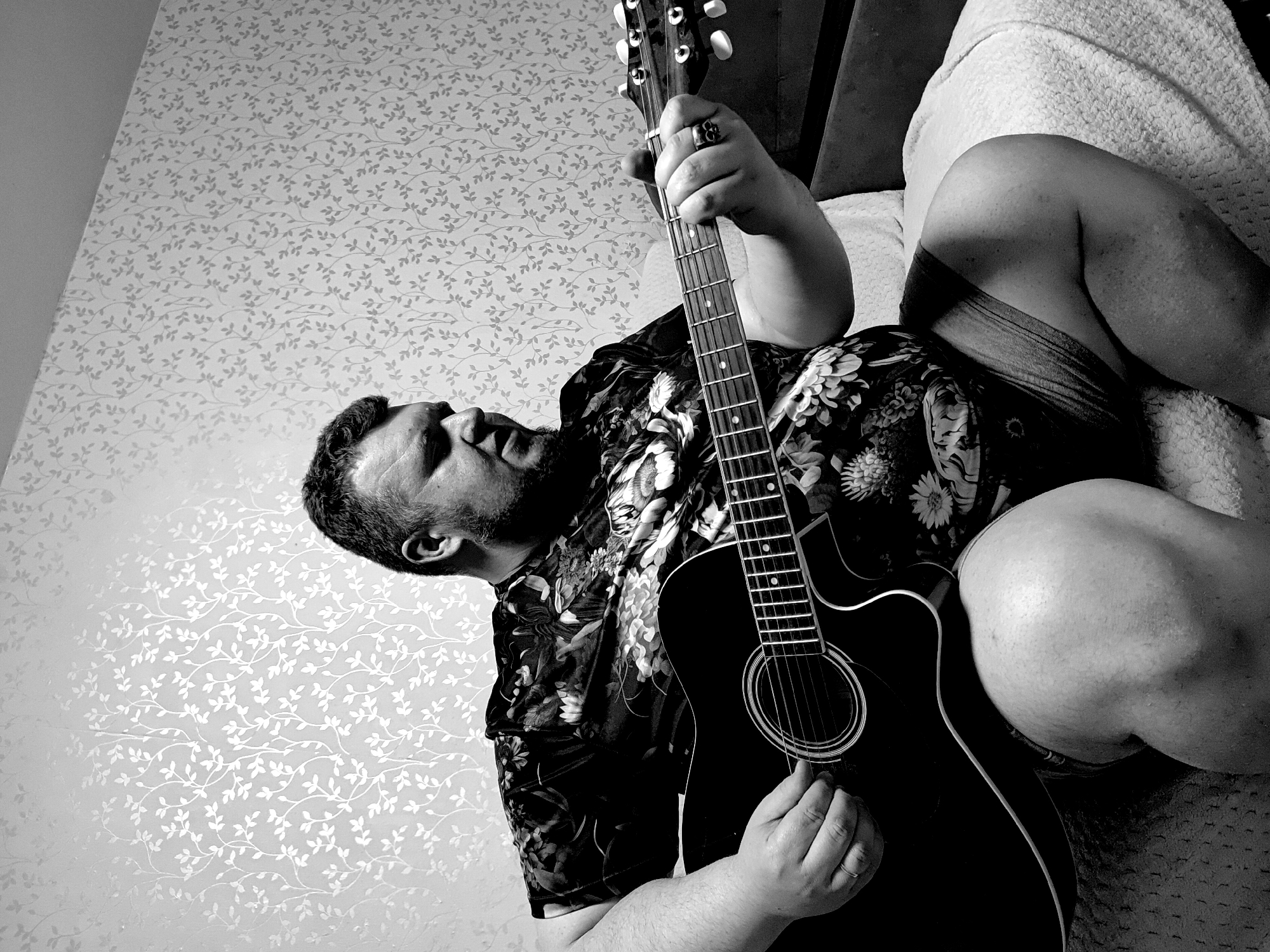 A black and white image captures a man lying down on his back, playing an acoustic guitar indoors. The man is positioned vertically in the frame, with his head towards the top left and his feet towards the bottom right. He has a beard and short hair, and his eyes appear to be looking upwards. He is wearing a dark, short-sleeved shirt with a dense floral or botanical pattern. A simple band ring is visible on his left ring finger, and a larger, possibly ornate ring (appearing skull-like or abstract) is on his right middle finger. His legs are partially visible, clad in dark shorts or underwear, resting on a textured, light-colored blanket or throw.
He is holding a dark-bodied acoustic guitar with light-colored binding and fret markers. His left hand is gripping the fretboard, while his right hand is positioned over the soundhole as if strumming. The background features a light-colored wallpaper with a repeating pattern of delicate, intertwining leaves or vines. The lighting creates a high-contrast scene, casting shadows and highlighting the textures of the wallpaper and the man's skin. The overall atmosphere suggests a moment of relaxed leisure and musical engagement in a home setting. There is no visible text. The location context is Марковский, Russia.