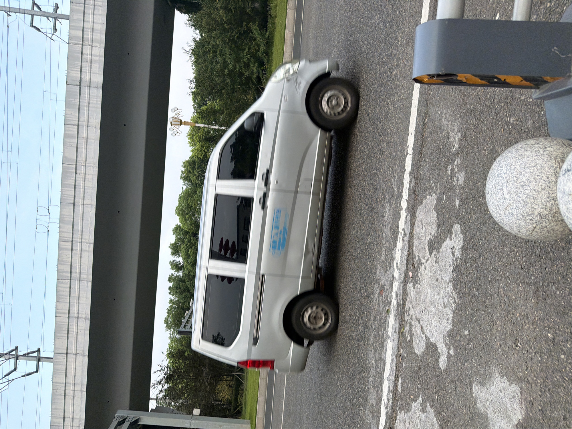 A silver van is captured in a side profile shot, traveling on a road beneath an elevated structure. The van has tinted windows and blue lettering on its side. The road surface is asphalt with white lane markings and some water stains. In the foreground, there are a couple of decorative spherical objects and a traffic barrier with yellow and black stripes. The background shows a line of green trees and utility poles with overhead wires against a pale sky. The scene is likely in the city of Jiangjin, China, given the context.