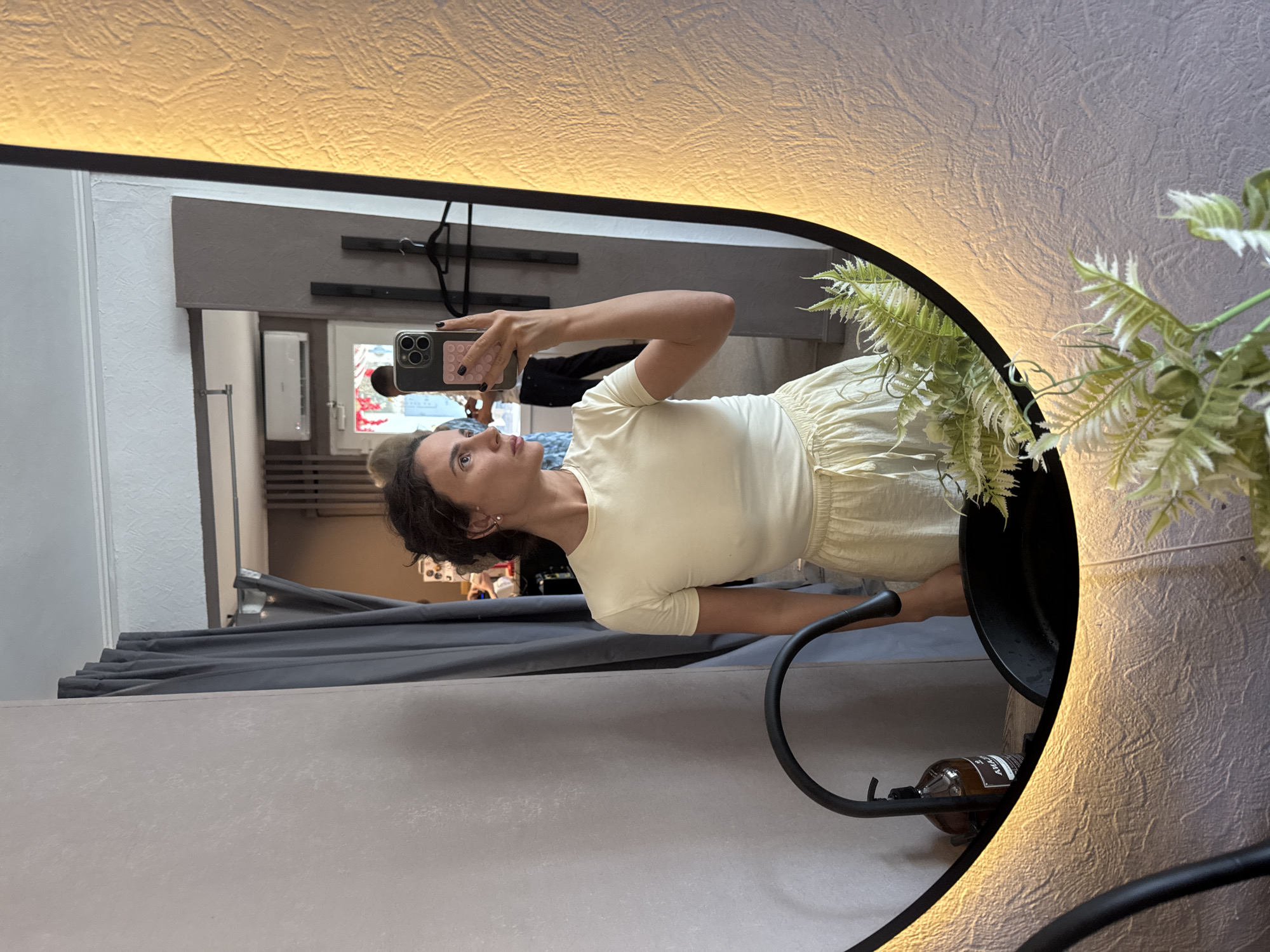 In a brightly lit room, a woman with dark, curly hair tied back takes a mirror selfie. She is wearing a light yellow t-shirt and matching shorts. Her reflection is visible in a large, oval mirror that is illuminated by a warm light from its border. In the background, through an open doorway, other people are visible, and there is an air conditioning unit mounted on the wall. The room appears to be a bathroom, with a black sink and faucet visible in the foreground of the mirror's reflection. The walls have a textured finish. The scene is devoid of any explicit indication of its location within Chaikovsky, Russia.