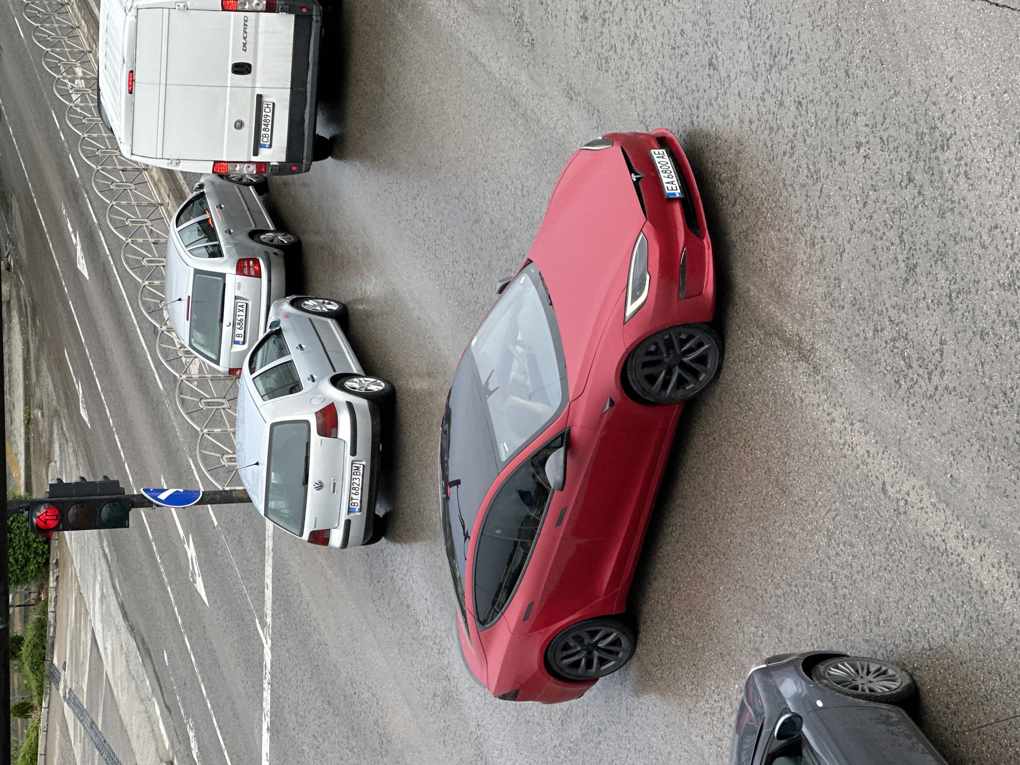 An elevated view captures a street scene in Veliko Tarnovo, Bulgaria, under what appears to be an overcast sky. The road surface is wet, suggesting recent rain or drizzle. Several vehicles are stopped, likely at a red traffic light, which is prominently illuminated on the left side of the frame.
In the upper part of the image, a white Fiat Ducato van is visible from the rear, displaying "DUCATO" and "FIAT" text, and the Bulgarian license plate "CB 8489 CH". Below it, two silver Volkswagen Golf cars are aligned. The rear Golf shows the license plate "B 6861 XA", while the one in front has "BT 6823 BM" and the "VOLKSWAGEN" emblem.
A striking red Tesla Model S dominates the center-right, angled slightly. It features a small "TESLA" logo and the Bulgarian license plate "EA 6800 AE" on its front bumper. In the bottom-right corner, part of a dark grey car is partially visible.
To the left, a traffic light displays a red signal. Next to it, a blue circular traffic sign with a white arrow pointing left indicates a mandatory left turn or turn ahead. White road markings, including arrows, are painted on the wet asphalt. A metal bike rack or barrier runs along the left edge of the street, bordering a sidewalk with some greenery visible at the bottom left. The image offers a factual depiction of urban traffic conditions.