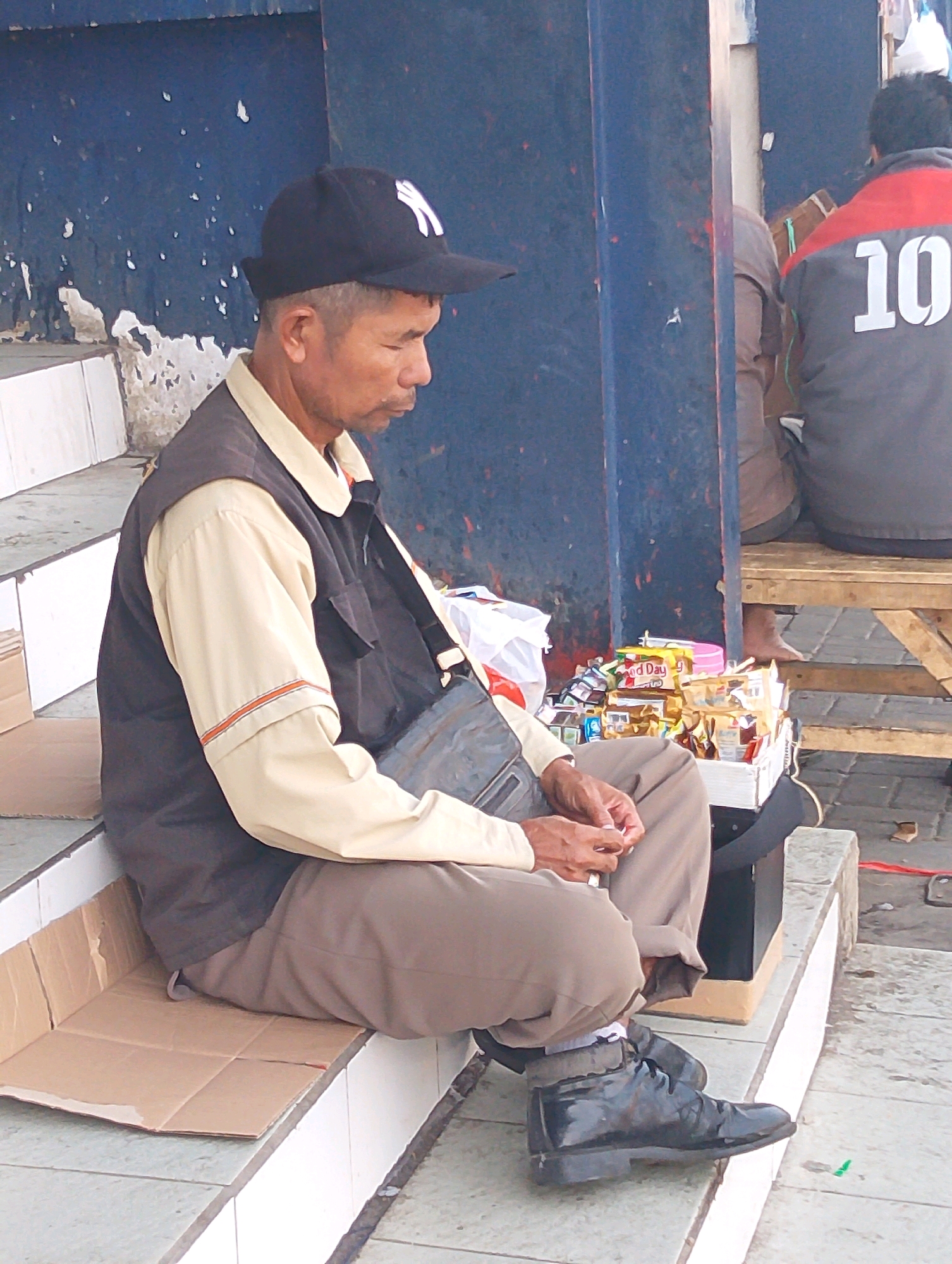 In Lembang, Indonesia, an elderly Asian man sits on a tiled outdoor staircase, covered with a piece of cardboard for comfort. He wears a black baseball cap with a white logo, a light-colored long-sleeved shirt with orange trim on the cuffs, and a dark vest over it. A dark bag is slung across his chest. His hands are clasped in his lap, and he appears to be looking down. Beside him, a small cart is filled with various packaged goods, including snacks and drinks. In the background, another person wearing a grey jacket with a large white "10" on the back is visible, seated on a wooden bench. The wall behind the man is painted dark blue with white speckles.