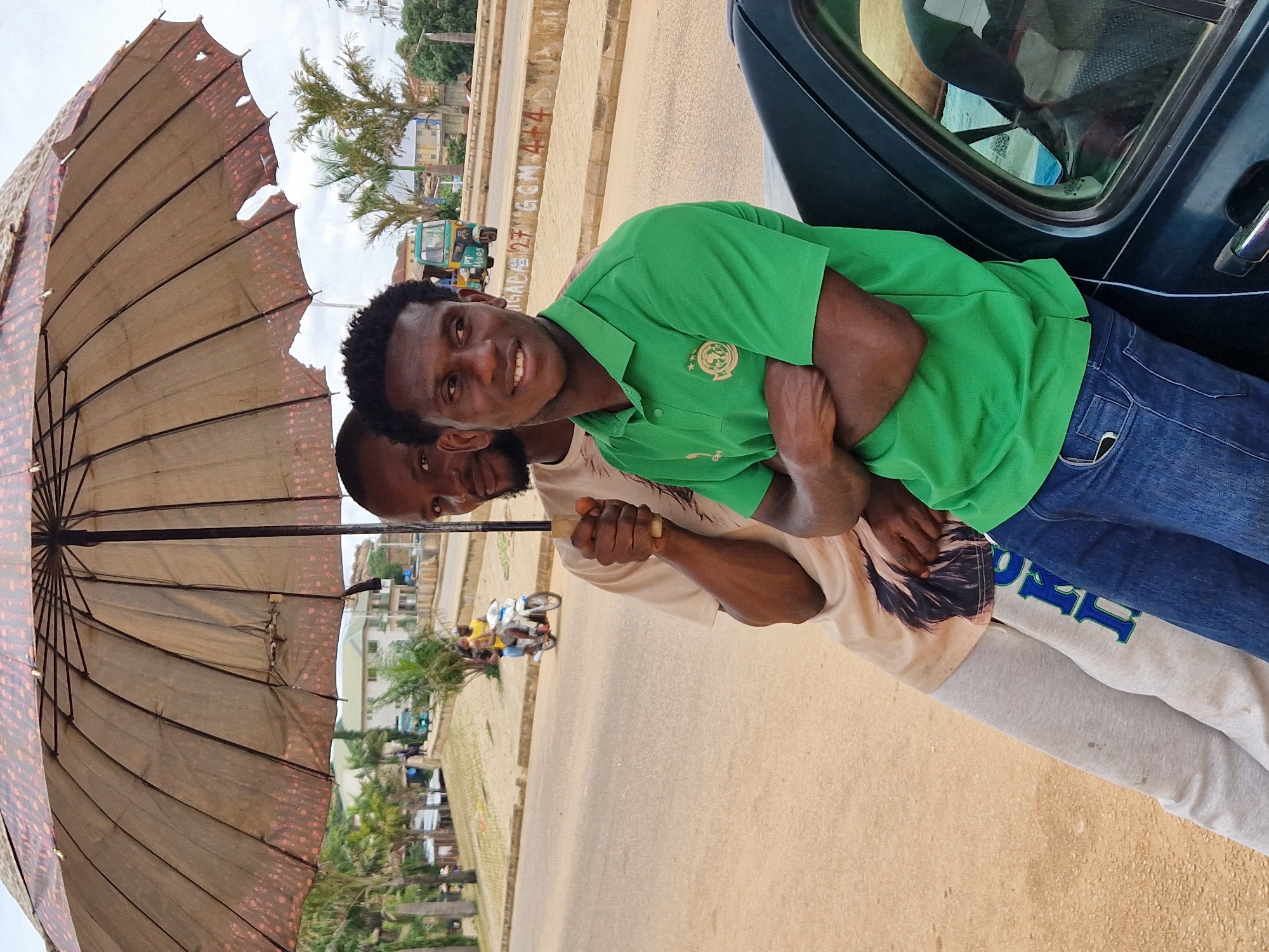 In Kabong, Nigeria, under a large, worn umbrella, two men stand by a dark-colored vehicle. The man in front, wearing a green polo shirt and blue jeans, has his arms crossed and is looking to the right with a smile. He is leaning against the car. The man behind him, partially visible and wearing a light-colored t-shirt and grey sweatpants, is holding the umbrella handle. The umbrella is patterned with red circles and has several holes. The background shows a street with buildings, palm trees, and a tricycle in the distance. The sky is overcast.