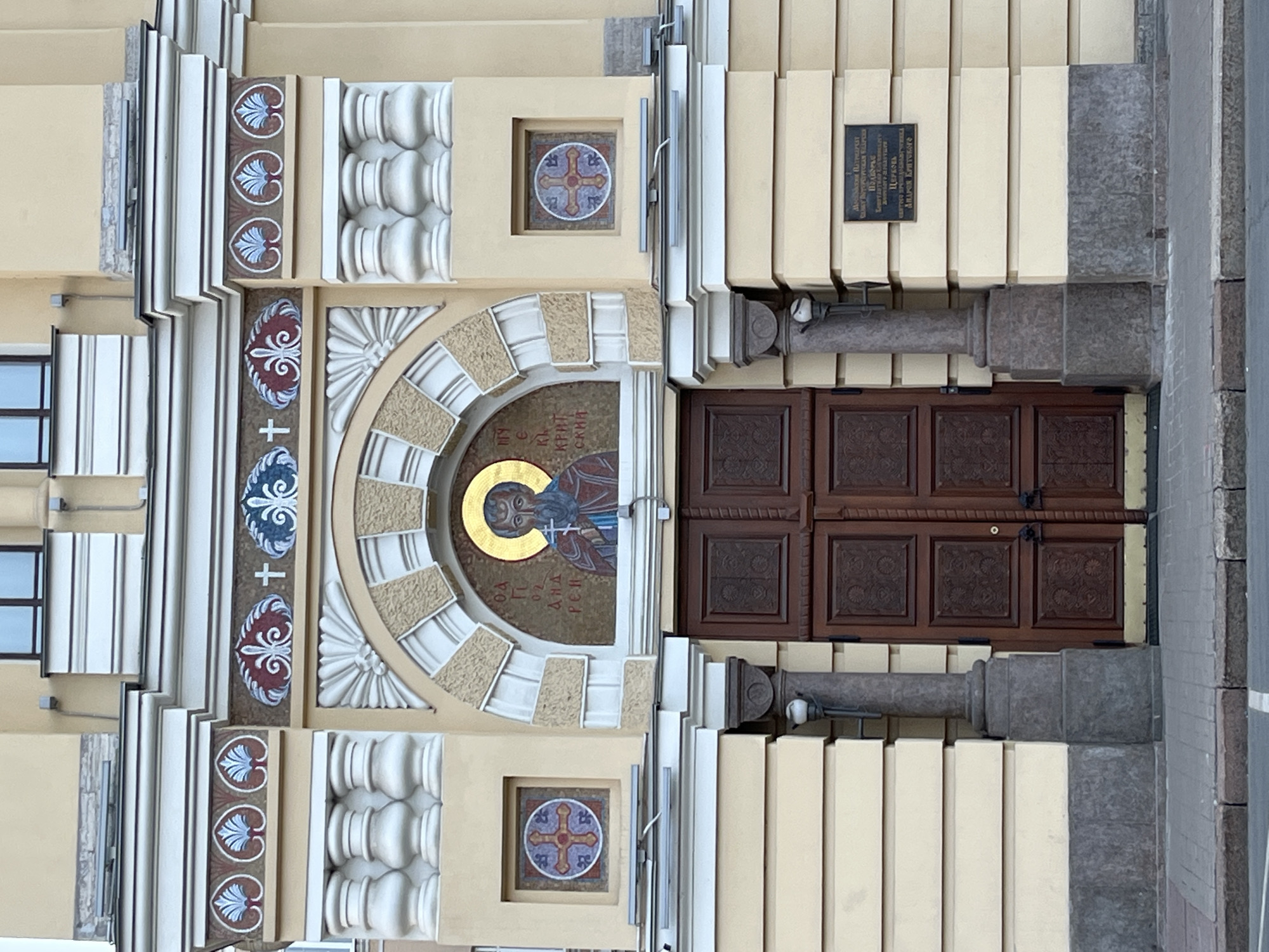 Here is a detailed description of the image:
**Scene:** The image depicts the entrance to a building.
**Architecture:**
The building's facade is decorated with intricate architectural details. The doorway features a rounded arch with a mosaic of a religious figure with a golden halo. Above the arch are decorative moldings, including rows of rounded elements, floral motifs, and patterns of leaves and hearts. Square tiles with a cross design are set into the wall above and below the arch. The doors are dark brown wood with square panels. A black plaque is mounted to the right of the door.
**Additional Information:**
Text on the plaque is partially readable.
**Environment:**
The image is taken outdoors, with a partial view of a street visible to the right.