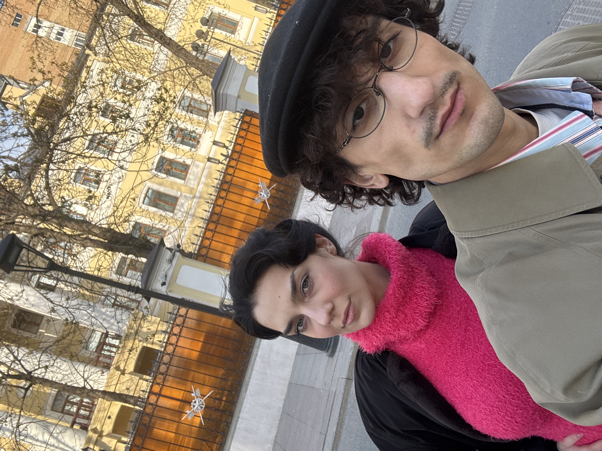 A selfie photograph, likely taken by the man in the foreground, features two individuals in an urban setting. The man, positioned slightly higher, wears a black beret and round eyeglasses. He has dark curly hair, a mustache, and a tan trench coat. Below him, a woman wears a bright pink fuzzy sweater and a black coat. The background shows a yellow building with multiple windows, leafless trees, and a decorative fence. Based on architectural style, the location is plausibly Moscow, Russia. The image exhibits an upward angle, typical of a selfie. The subjects appear to be posing for the photograph.