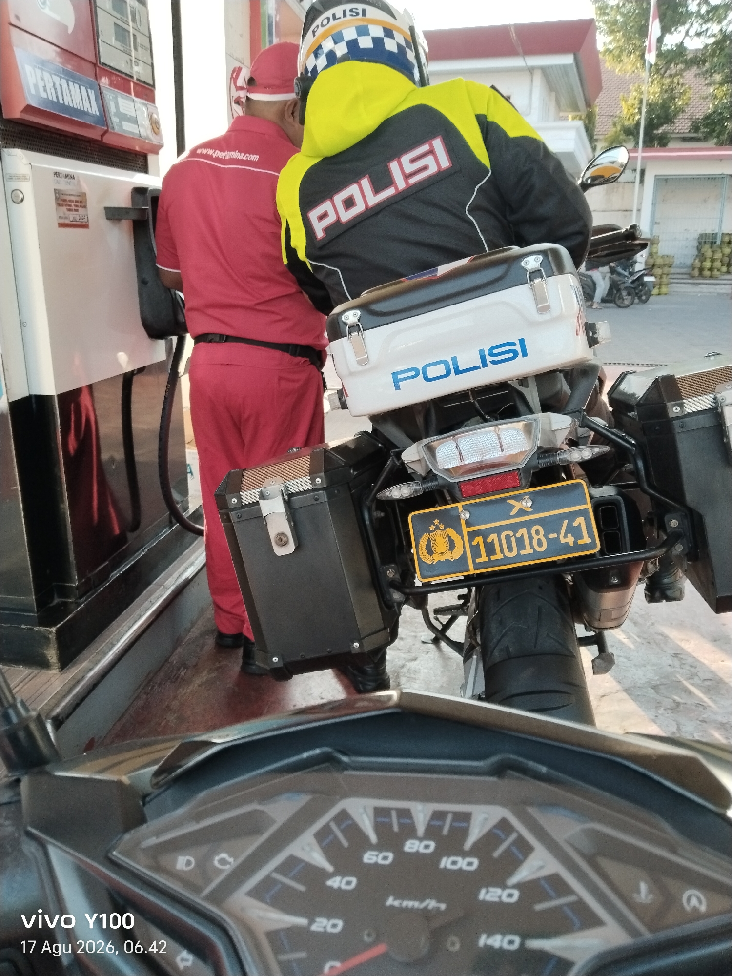 The image shows a police officer on a motorcycle at a Pertamina gas station in Patokan, Indonesia. The officer is wearing a yellow and black jacket with "POLISI" written on it, and a blue and white helmet. The motorcycle is equipped with saddlebags and a police insignia on the license plate. A gas attendant in a red uniform is standing next to the motorcycle, appearing to be assisting the officer. The gas station has several pumps and the background suggests an outdoor environment with some buildings and trees. The timestamp on the image indicates it was taken on August 17, 2026, at 06:42 AM.