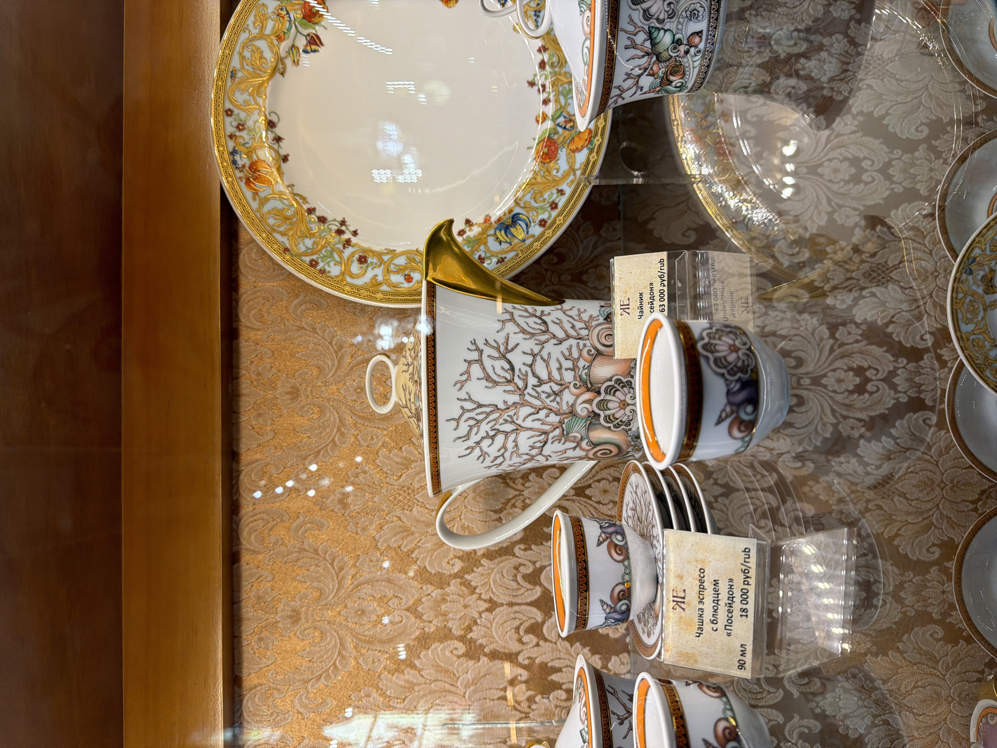 This image captures a display of exquisite porcelain tableware, likely in a retail environment, within Saint Petersburg, Russia. The various items are showcased on a clear glass shelf, creating visible reflections of overhead lighting. In the background, there is a decorative golden-brown patterned fabric or wallpaper.
The subjects include a large, ornate plate, a teapot, and several espresso cups with saucers. The large plate, positioned towards the top, features a sophisticated design with a broad rim decorated with rich gold scrollwork, pale blue details, and colorful floral motifs in shades of red, orange, and blue.
The teapot and cups exhibit a cohesive marine-themed pattern, characterized by intricate branching coral structures in earthy tones, alongside various stylized seashells and sea creatures, rendered in greens, blues, and oranges. The interior of the cups is a vibrant orange, contrasting with the detailed exterior. The teapot stands out with its elegant, upward-curving golden handle. The espresso cups are presented with their matching saucers, some stacked neatly.
Visible text on price tags provides specific details. One tag identifies an "Espresso cup with saucer 'Poseidon'" (Чашка эспресо с блюдцем «Посейдон»), noting its "90 ml" (90 мл) capacity and a price of "18,000 rubles/rub" (18 000 руб/rub). Another tag points to a "Teapot 'Poseidon'" (Чайник «Посейдон») priced at "63,000 rubles/rub" (63 000 руб/rub). The Russian text and currency confirm the provided location context. The scene depicts a static product display, highlighting the artistry and value of the luxury tableware.