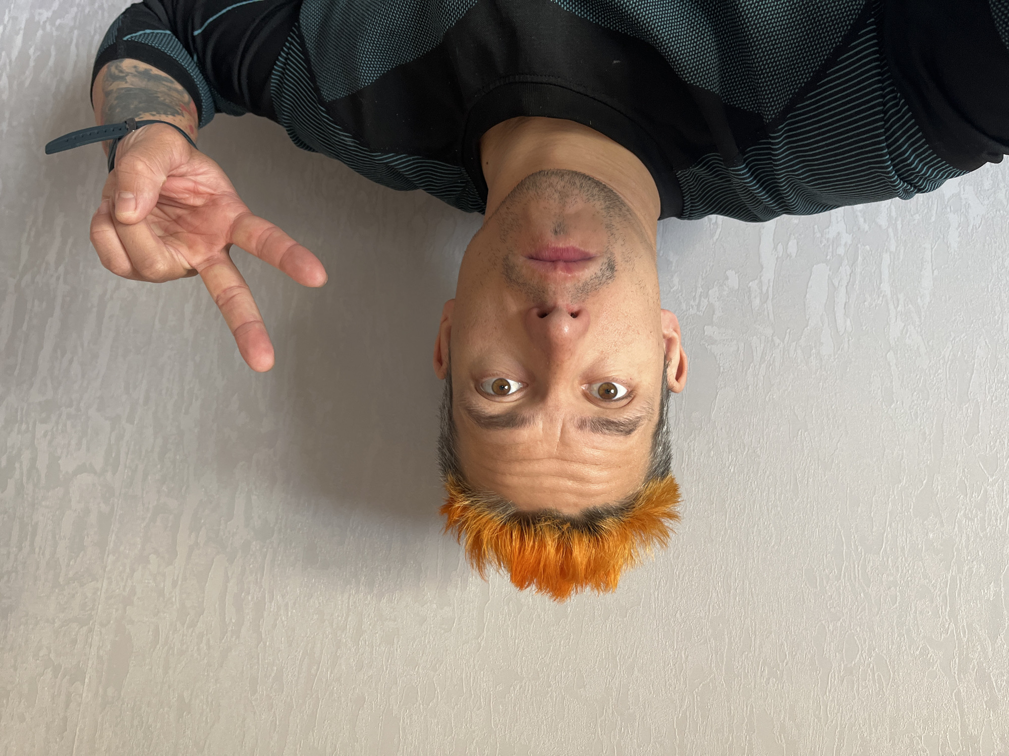A person is upside down against a light-colored, textured wall. The person has tan skin, brown eyes, and a trimmed beard. Their hair is short and dyed orange at the front. They are wearing a black shirt with teal geometric patterns. The person is making a peace sign with their left hand, which has a tattoo on the forearm and a watch on the wrist. The specific location within Chaikovsky, Russia, is not discernible from the image.