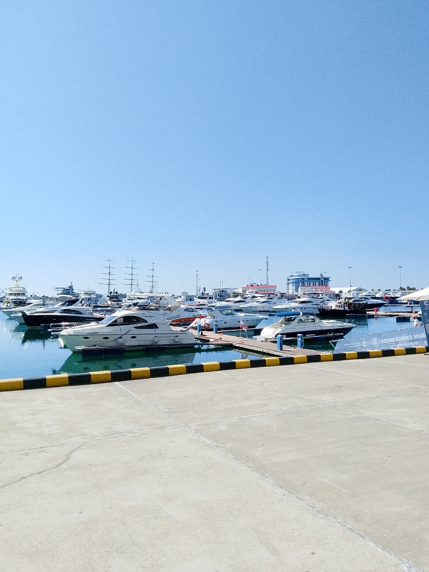 The image depicts a marina filled with various yachts and boats docked under a clear blue sky. In the foreground, a concrete pier with yellow and black striped bollards lines the edge of the water. The water itself is calm and reflects the boats and the sky. Several white yachts of different sizes are prominently featured, some with sleek, modern designs. In the background, larger sailing vessels with multiple masts are visible, along with other boats and structures of the marina, including a blue building with red accents. A sign partially visible reads "SOCHI GRAND MAR" suggesting the location as Sochi Grand Marina. There are no people clearly visible interacting with the boats or the environment. The overall scene conveys a sense of leisure and maritime activity.