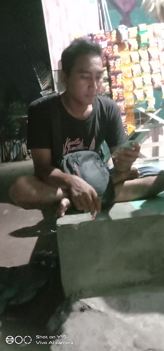 In Jabung Sisir, Indonesia, a single male individual is depicted at night, squatting or leaning on a low concrete ledge. He wears a dark, short-sleeved t-shirt with a light graphic and dark shorts, and is barefoot. A black sling bag rests across his chest. In his left hand, he holds and views a light-colored smartphone. His right hand holds a lit cigarette with a visible ember, positioned over the concrete ledge. The background immediately to his right features a brightly illuminated vertical display rack showcasing numerous packaged snack items against a light-colored wall, indicating a vendor stall. The area to his left is dark and indistinct.
