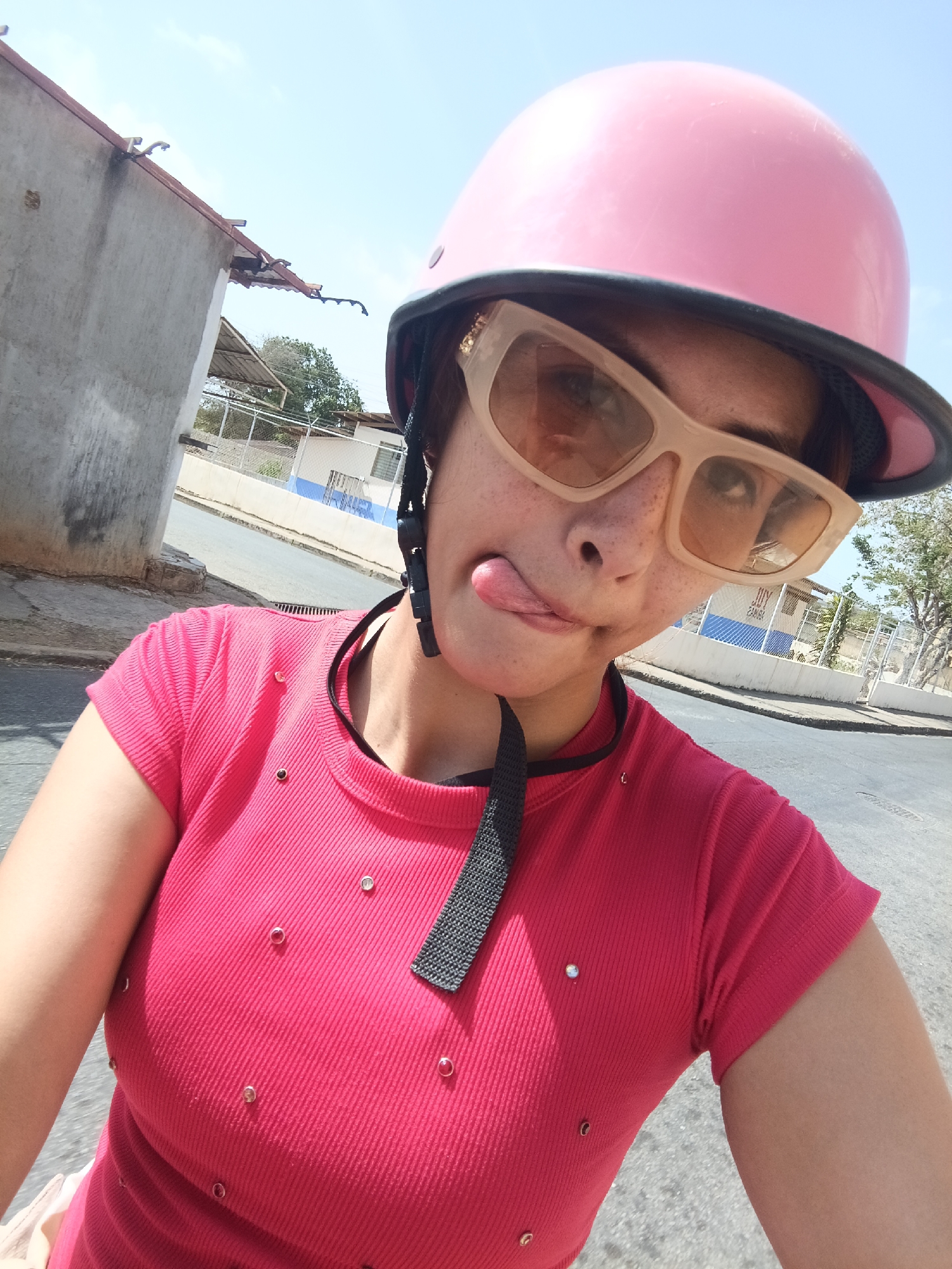 A medium close-up selfie shows a woman wearing a pink helmet, beige sunglasses, and a red t-shirt with metal studs, sticking her tongue out. She is positioned on a paved street in Juan Griego, Venezuela. The background includes a white building with blue accents, a chain-link fence, trees, and other buildings. The sky is partly sunny.