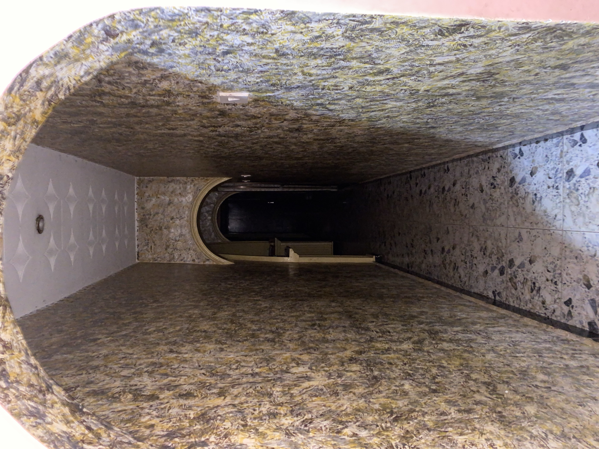 The image shows a narrow indoor hallway with patterned wallpaper and a tiled floor. The hallway is arched at the entrance and has another arched doorway at the far end, which is dark and mostly obscured. The wallpaper has a mottled pattern of brown, yellow, and grey. The floor is tiled with a speckled pattern of grey and white. There is a light switch visible on the right-hand wall. The scene is consistent with an interior space in Jos, Nigeria, likely a residential or commercial building.
