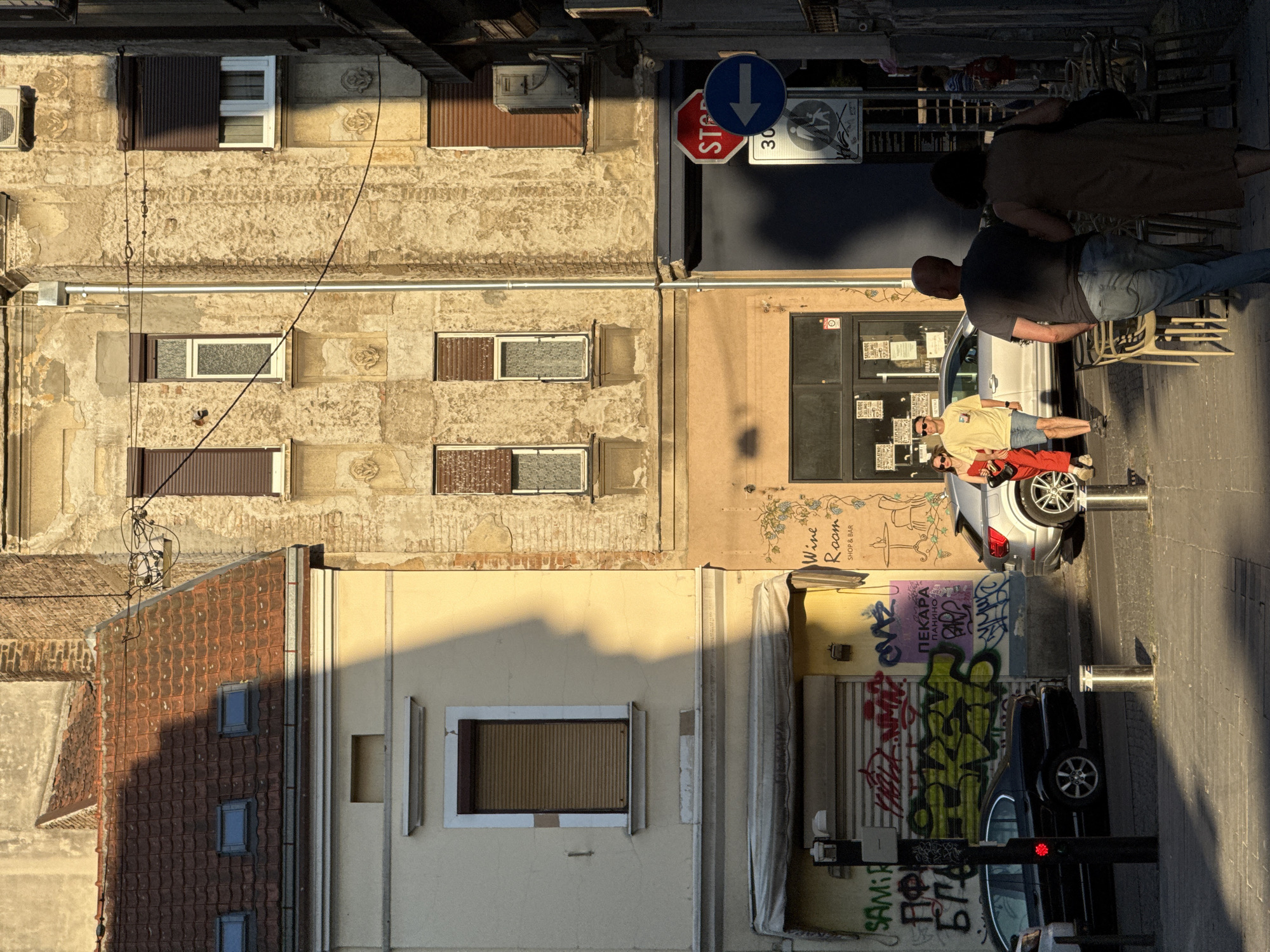 The image depicts a street scene in Belgrade, Serbia, bathed in the warm, golden light of what appears to be late afternoon or early morning. Two buildings dominate the frame. The left building is lighter in color with a red tiled roof and dormer windows, while the building to its right is older, with crumbling yellow stucco, exposed brickwork, and multiple windows with brown rolling shutters.
In the foreground, a black sedan is parked on the left side of the street, its rear end visible. To the right of the sedan, a modern silver SUV is parked, and two couples are walking along the sidewalk. The first couple, a man in a yellow t-shirt and blue shorts and a woman in a red jumpsuit, are walking towards the SUV. The second couple, a man in a grey t-shirt and jeans and a woman in a brown dress, are walking away from the viewer towards the right side of the frame.
Graffiti covers the lower section of the building on the left, with various tags and markings in different colors, including prominent green and purple writing. A street sign with a red stop symbol and a blue arrow indicating a left turn, along with a speed limit sign of "30" and a pedestrian crossing symbol, are visible on the right side of the street.
The street is paved with cobblestones, and metal bollards are placed along the sidewalk. Outdoor cafe seating, with empty chairs, is visible on the right side of the frame, suggesting commercial activity. The overall impression is one of an urban environment with a mix of older and newer architecture, residential and commercial spaces, and everyday life unfolding on the street.