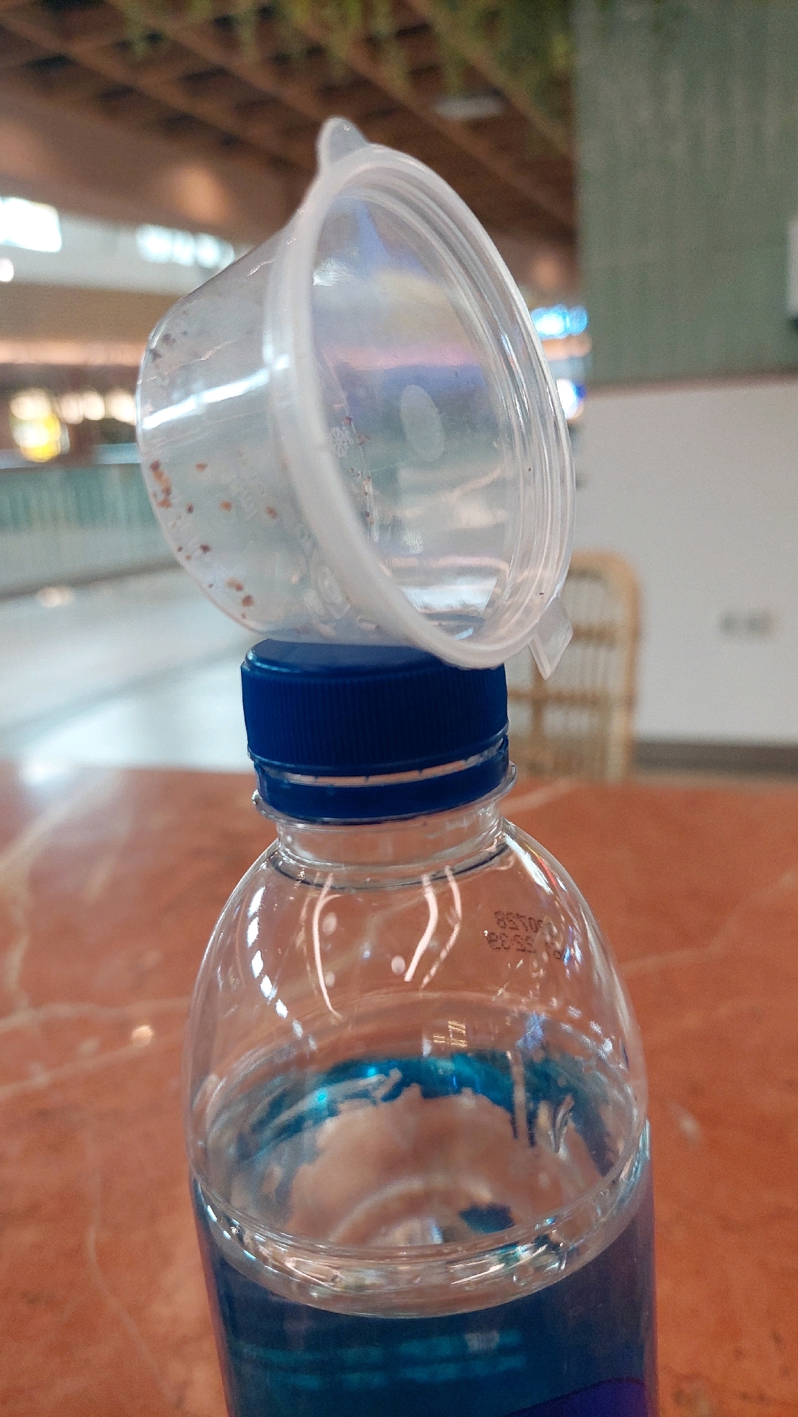 A plastic water bottle with a blue cap and some blue liquid inside is placed on an orange table. A clear plastic lid, with some residue inside, is balanced on top of the bottle cap. The background suggests an indoor setting, possibly a mall or restaurant, with wooden ceiling beams and a blurred view of the environment. The image is taken from a medium close-up perspective. No people are visible in the image. The scene is forensic and objective, devoid of any subjective interpretation or emotional content. The location is North Jakarta, Indonesia.