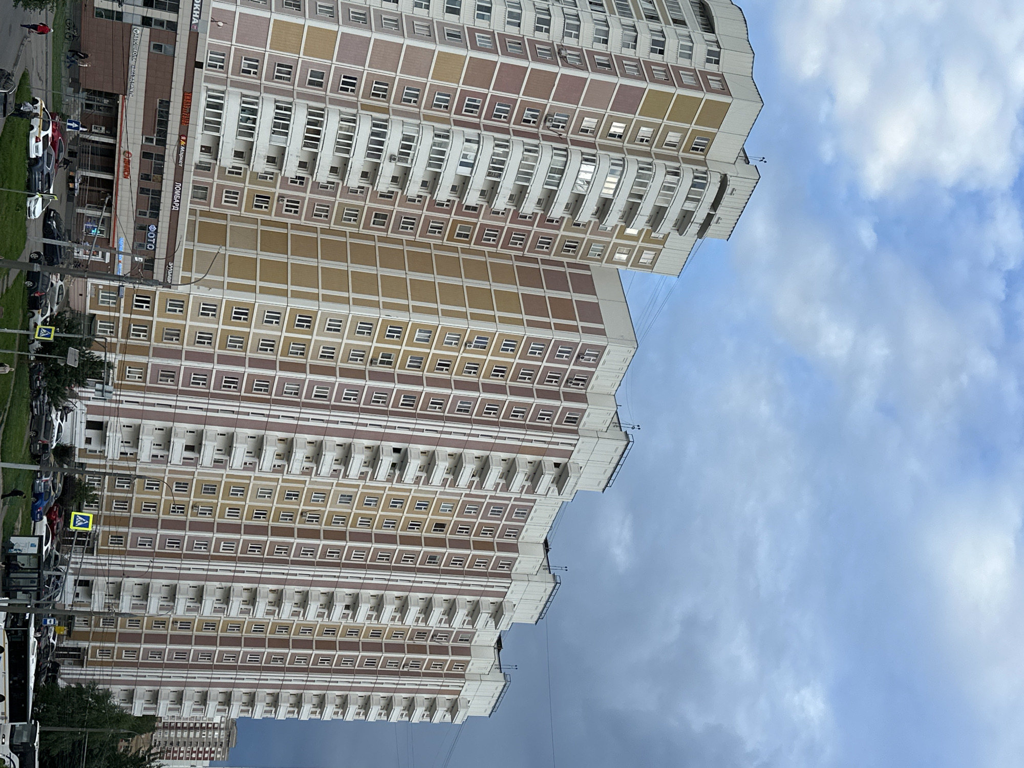 The image depicts a view of several multi-story apartment buildings in Podolsk, Russia. The buildings are tall, with multiple balconies and windows. The facades are a mix of light colors, including white, beige, and pinkish-brown. The sky is partly cloudy with a mix of blue sky and darker, more ominous clouds towards the right side of the frame. In the foreground, there are streets with cars parked and driving, as well as some pedestrians. Commercial establishments with signs are visible on the ground floors of some buildings. One sign reads "Я Маркет," indicating a supermarket. Another sign says "ФОТО," suggesting a photo shop. The overall scene captures a residential and commercial area on an overcast day in Podolsk.