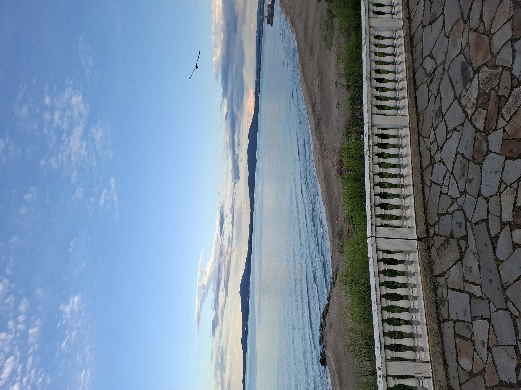 The image depicts a coastal scene in Burgas, Bulgaria. The perspective is from an elevated position, likely a balcony or terrace, as indicated by the white balustrade on the right side of the frame and the stone-tiled floor. Looking out, there's a sandy beach meeting a body of water, presumably the Black Sea. The water is relatively calm, with gentle waves visible. The background features a distant landmass or coastline, partially obscured by clouds and haze. The sky is predominantly blue, with some scattered clouds. A bird is flying in the sky. The scene suggests a serene and tranquil environment.