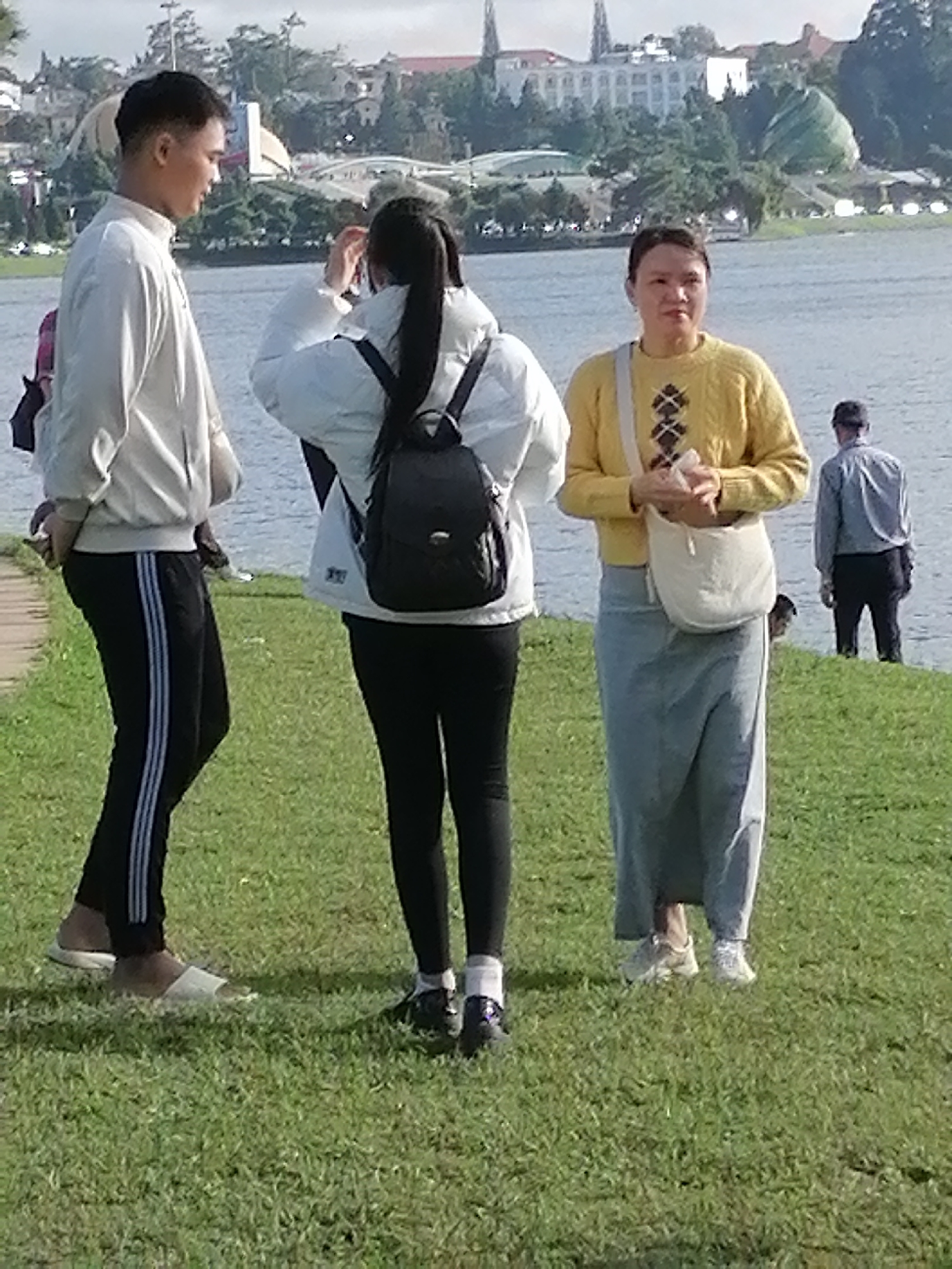 A group of people stand on a grassy bank overlooking a body of water. The scene is set against a backdrop of buildings and trees in the city of Da Lat, Vietnam. In the foreground, a young man in a white jacket and black athletic pants stands to the left. Next to him, a young woman in a white jacket and black pants is seen from behind, with her hair in a ponytail and a black backpack. To her right, a woman in a yellow sweater and a long grey skirt holds a white bag. Further in the background, a man in a light blue shirt and dark pants stands near the water's edge. The weather appears to be sunny with some clouds.