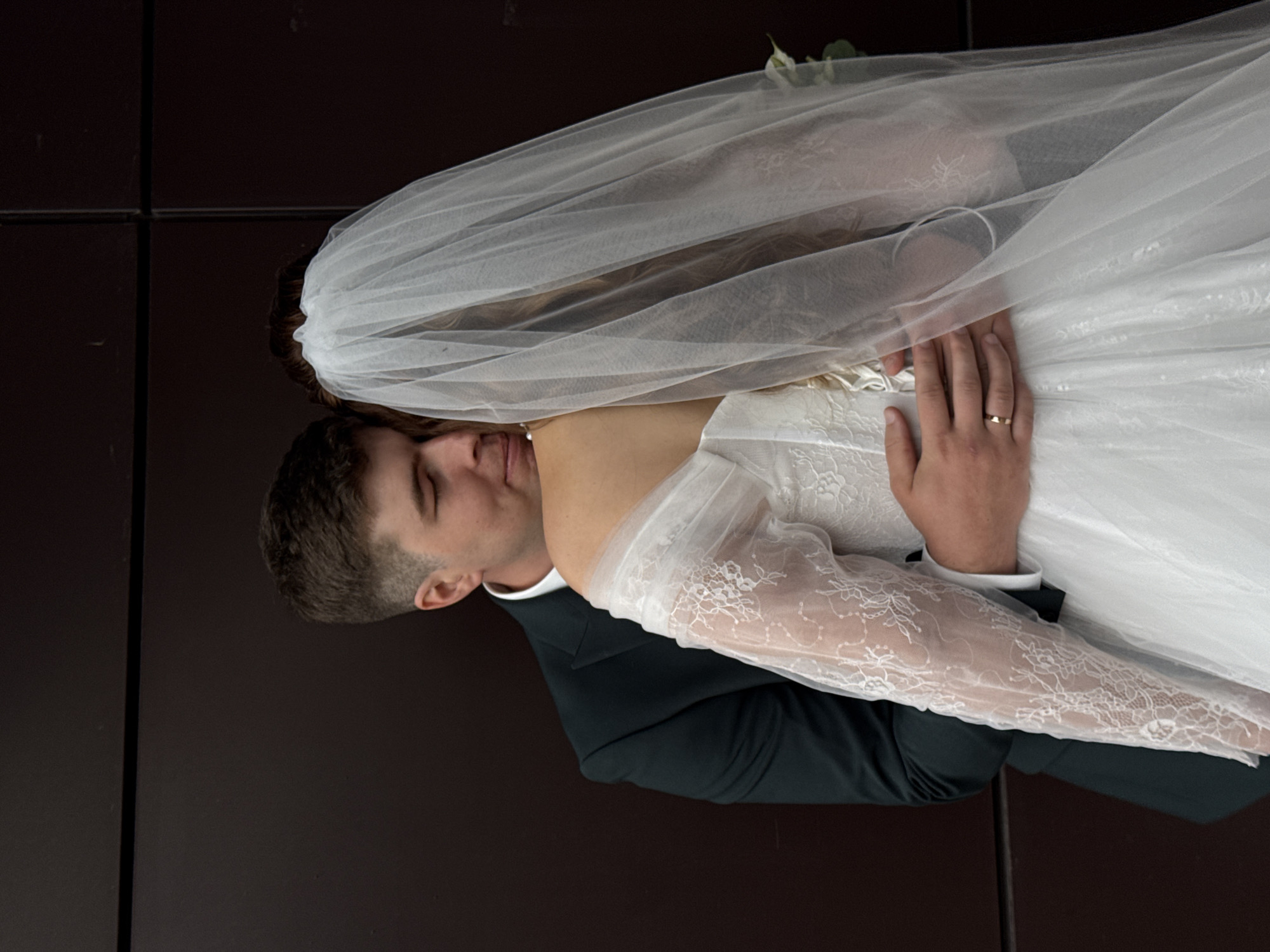 The image captures a tender embrace between a bride and groom, likely during their wedding or a related photoshoot, set against a minimalist, dark brown background. The bride, positioned on the right side of the frame, is wearing an elegant white wedding dress featuring delicate lace sleeves and a flowing sheer veil that drapes over her head and shoulders. Her light-colored hair is visible through the veil. Her left hand, adorned with a simple ring on her ring finger, is gently wrapped around the groom's back. The groom, situated on the left, is dressed in a dark suit with a white shirt and has short, dark hair. He is seen resting his head affectionately against the bride's shoulder and neck. Both appear to have their eyes closed or looking downwards, creating an intimate and emotional atmosphere that conveys love and serenity. The plain, dark backdrop ensures that the focus remains entirely on the couple and their heartfelt connection. No specific time of day, weather conditions, or text are visible within the frame. This intimate moment is situated in Chaikovsky, Russia.