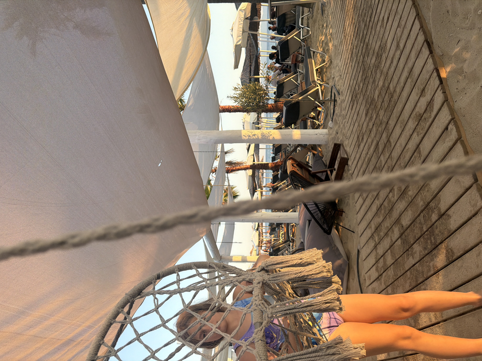 The image depicts a person lounging in a hanging chair on a wooden boardwalk at what appears to be a beachside establishment. The subject, a young female, is partially visible in the hanging chair, wearing a colorful swimsuit. Her legs extend towards the right side of the frame, resting on the wooden planks.
The setting is a beach with a boardwalk made of weathered wooden planks. In the background, there are rows of lounge chairs and umbrellas, suggesting a place for relaxation and sunbathing. Palm trees and smaller olive trees are interspersed throughout the scene, adding to the tropical or Mediterranean ambiance. Beyond the beach furniture, a body of water, likely the sea, is visible under a clear sky.
The lighting suggests it is either early morning or late afternoon, with a warm, golden light casting long shadows. The sky is bright and clear, indicating fair weather. The overall mood is one of leisure and relaxation, enhanced by the tranquil setting and the golden hour lighting.
There is no visible text in the image. The focus is on the serene beach environment and the relaxed posture of the person in the hanging chair.