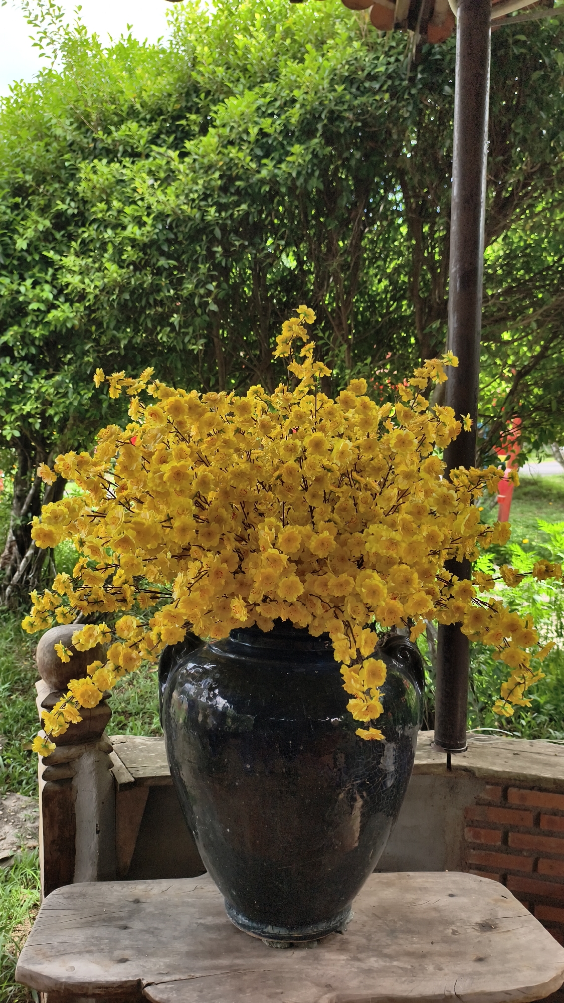 A large, dark blue ceramic vase filled with bright yellow artificial flowers sits on a weathered wooden table. The vase is positioned outdoors, with lush green trees and foliage in the background. A dark, cylindrical pole is visible to the right of the vase. The overall scene suggests an outdoor decorative arrangement, likely in a garden or patio setting, with an emphasis on the vibrant floral display.