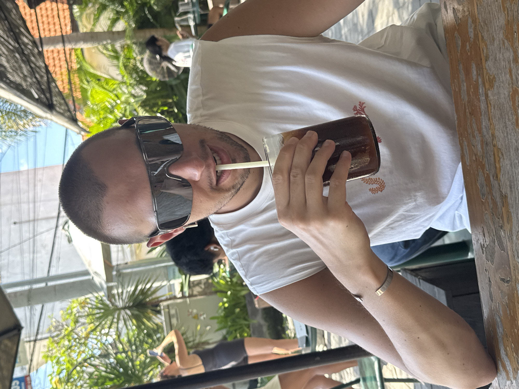 A man with short, dark hair and wearing sunglasses is drinking from a glass with a straw. He is wearing a white t-shirt and a silver bracelet. The setting appears to be outdoors, with vegetation visible in the background. There are glimpses of other people in the scene. The location is North Kuta, Indonesia.