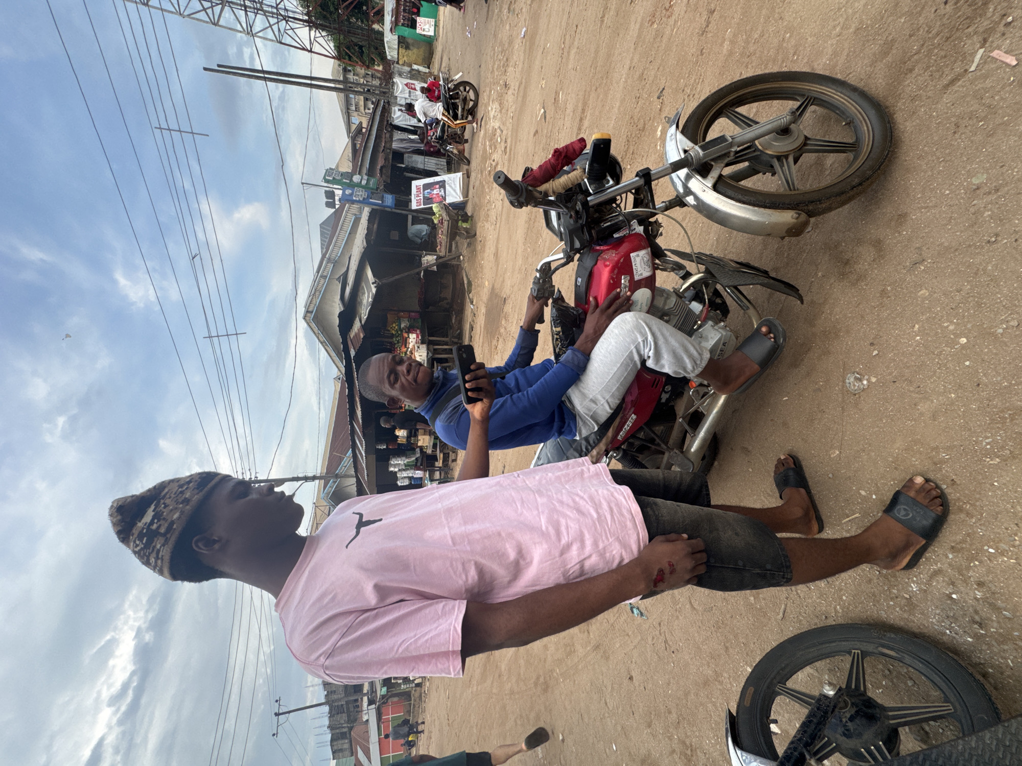 In Ihiagwa, Nigeria, two young men are interacting on a dusty street. The man on the left, wearing a pink t-shirt and a patterned hat, stands with his left hand on his hip, displaying an injury with dried blood on his hand. The man on the right is seated on a red motorcycle, wearing a blue long-sleeved shirt and grey pants. He is holding a smartphone in his right hand, seemingly showing it to the standing man. The motorcycle is parked on a dirt road, with another motorcycle visible in the foreground on the left. In the background, market stalls with corrugated iron roofs line the street, suggesting a commercial area. Overhead, power lines crisscross the sky, and a tall metal tower is visible on the right. The environment is characteristic of a busy Nigerian town.