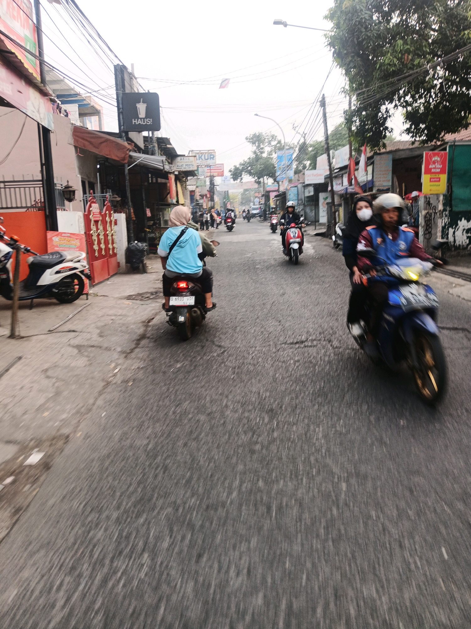 The image depicts a street scene in Bandung City, Indonesia, characterized by a slightly overcast sky and a blurred, motion-effected perspective. The street is lined with buildings, some of which display signage. On the left side of the image, a white and blue scooter is parked near a red metal gate. Further down the street, a person wearing a light blue t-shirt and a hijab is riding a scooter. In the middle ground, two individuals on a red scooter are visible, and to their right, two people on a blue scooter are also in motion. The road surface appears to be asphalt, and there are overhead power lines and streetlights present. The overall impression is one of a bustling urban environment captured during daylight hours.