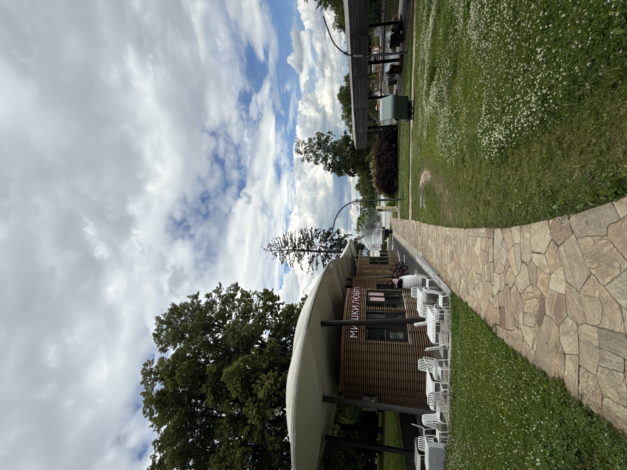 The image depicts a park scene in Moscow, Russia, with a modern cafe structure in the foreground. The cafe has a slatted wooden facade and a white, curved canopy. Outdoor tables and chairs are arranged in front of the cafe on a paved area. A stone pathway leads away from the cafe and into a grassy area dotted with small white flowers, possibly clover. In the background, trees and a body of water with a fountain can be seen. The sky is cloudy with patches of blue. A few people are visible near the cafe and in the distance. The overall impression is of a pleasant, possibly recreational, outdoor space.