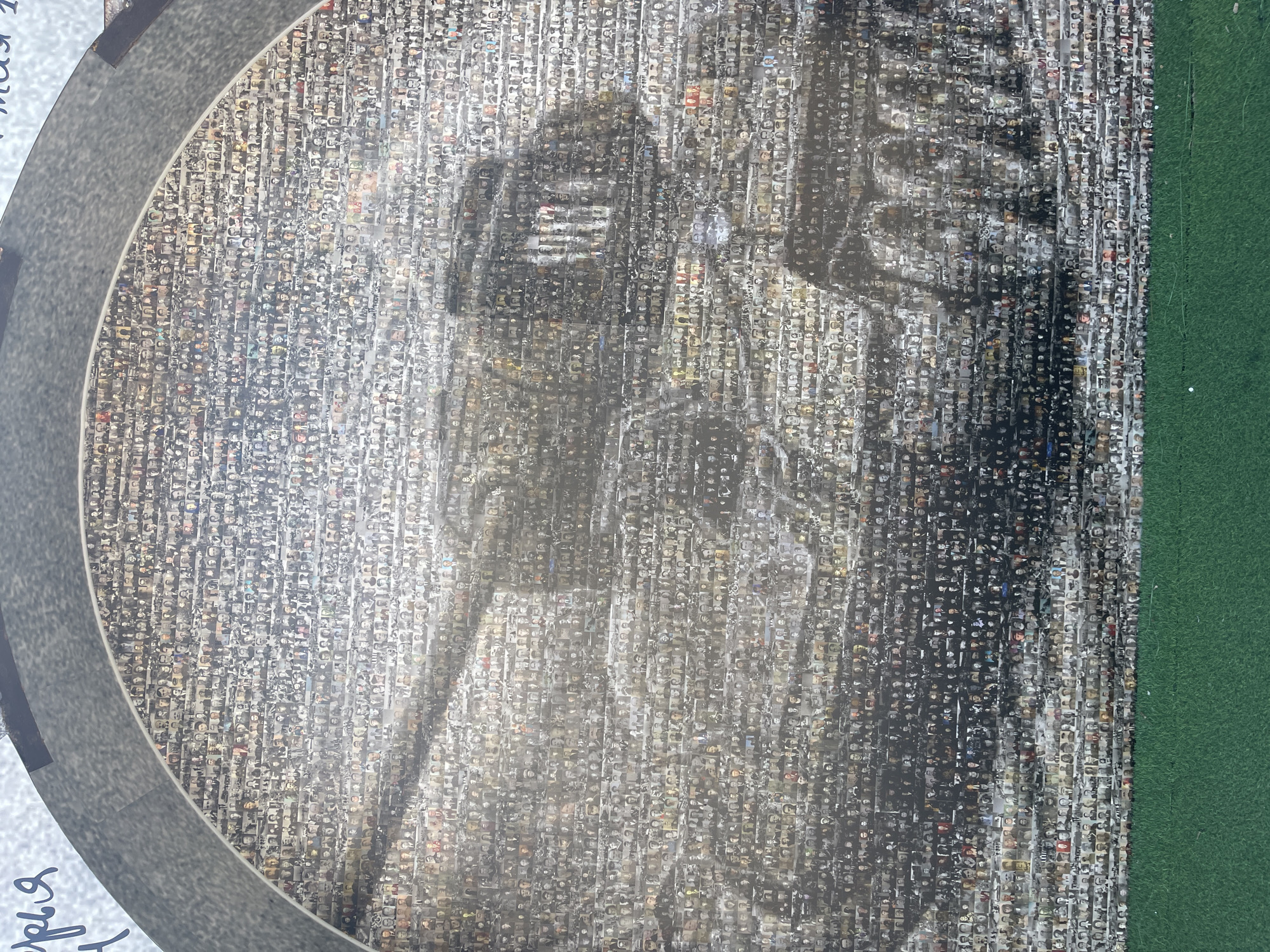 The image is a close-up of a mosaic created from numerous small photographs of people's faces. These photographs are arranged in vertical columns, forming a larger image which appears to be a silhouette of a person, possibly standing with a staff or long object. The mosaic is circular in shape, bordered by a grey, textured material on the left and a green border on the right. The overall tone of the mosaic is monochromatic, with shades of grey, black, and white, but some of the individual photographs contain hints of color.
The subjects are predominantly people, featured in individual portraits that make up the mosaic. The larger image formed by these portraits suggests a scene or a representation of an individual, but the detail is obscured by the nature of the mosaic.
There is no discernible activity or event taking place in the mosaic itself. The arrangement of faces implies a collection or commemoration of individuals.
Notable details include the circular framing of the mosaic, the contrast between the textured grey border and the smooth green border. The lighting appears to be even, with no strong shadows or indications of time of day. The location is indicated as Zelenogradsk, Russia, though no specific landmarks are visible within the mosaic.
Visible text includes handwritten script in a light blue color on the lower left side of the image, with the beginning of a word visible: "яря". There is also faint text on the upper part of the image, also in blue script, that appears to read "мая 2". The rest of the mosaic is composed of the small photographs of faces, making individual text recognition impossible.