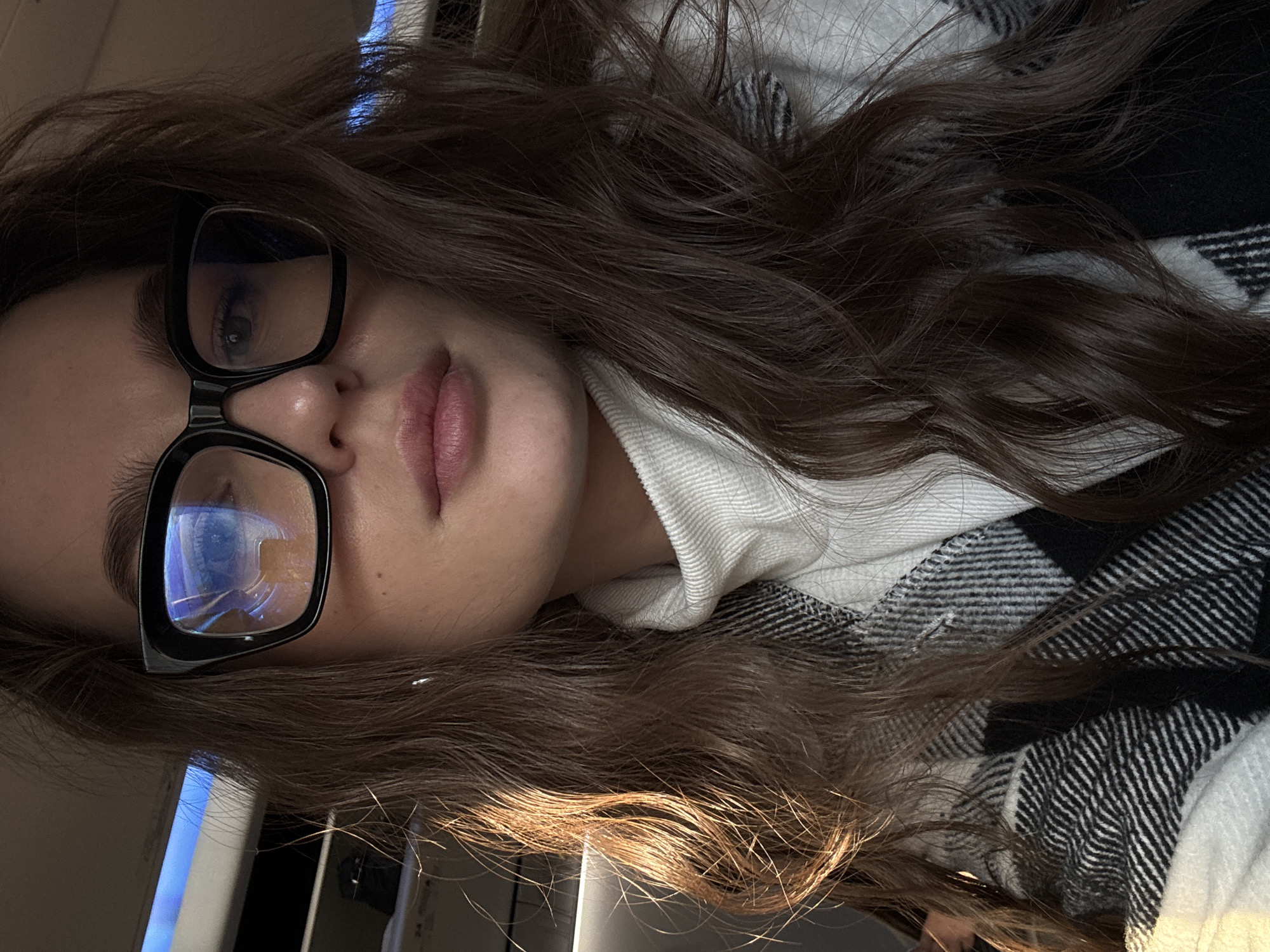 The image shows a woman with long, wavy brown hair wearing black-framed glasses. She is wearing a white turtleneck sweater and a black and white plaid flannel shirt. The background appears to be indoors. There is no information available to confirm the location is in Новое Мартьяново, Russia.
