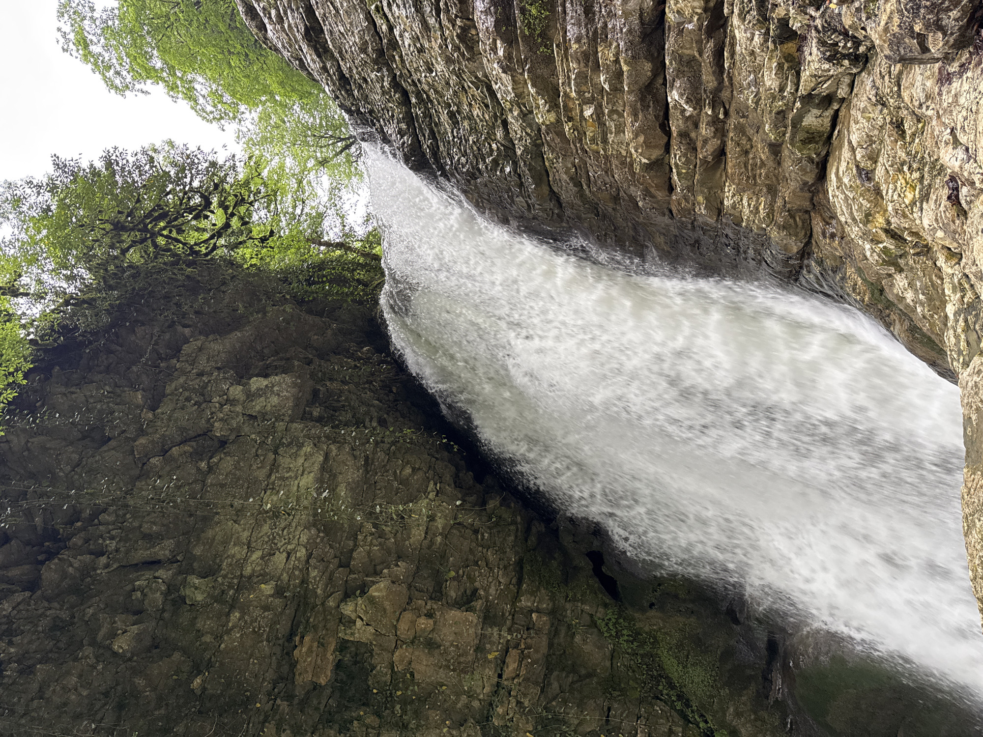 A photograph captures a waterfall cascading down a rugged cliff face. The water appears as a blurred stream, indicating movement. The cliff on the right side of the frame displays a stratified rock formation, with vertical layers and varying textures. Vegetation, including trees and moss, grows on the left cliff face, adding a touch of green to the scene. Above, the sky is visible through the opening between the cliffs, appearing bright and overcast. This image is most likely taken in Tsabal, Abkhazia.