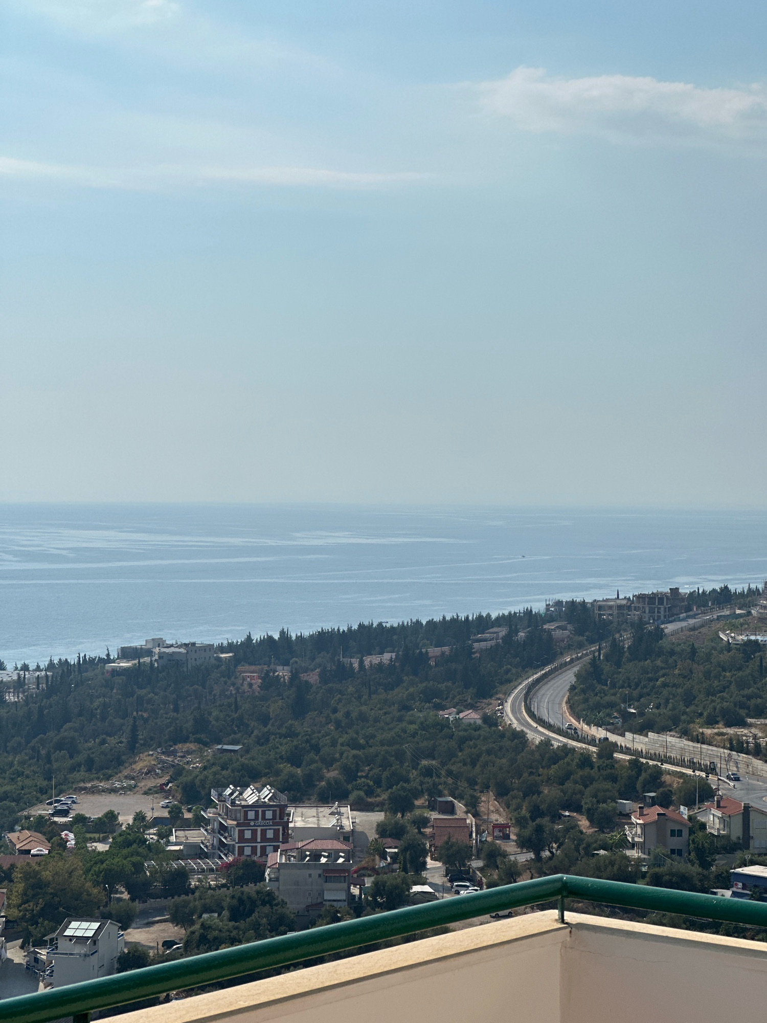 The image is a wide shot of a coastal town or city overlooking the sea. In the foreground, a white balcony railing with a green metal handrail is visible. The middle ground is filled with dense green trees covering rolling hills. Scattered among the trees are various buildings, including houses with red roofs, a larger building with the word "GRECCIA" written on it and solar panels on its roof, and some structures under construction. A curved road with cars can be seen winding through the hills. In the background, the calm, blue sea stretches to the horizon, meeting a pale blue sky with a few wispy clouds. The overall impression is of a sunny day in a developed coastal area.