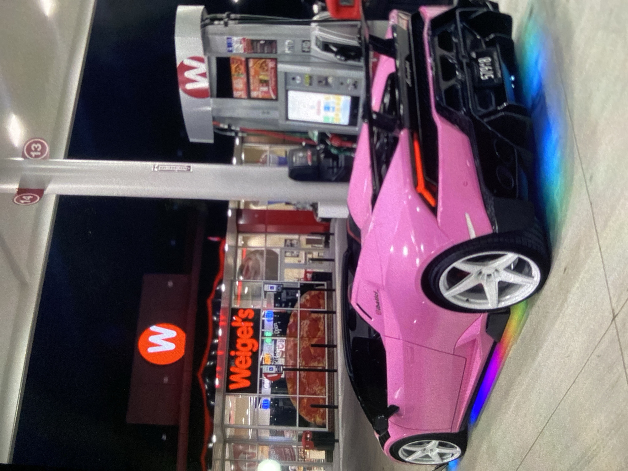 A pink Lamborghini is parked at a Weigel's gas station. The gas station has a red and white "W" logo and the name "Weigel's" in red neon letters. A gas pump is visible next to the car. The license plate on the car reads "5T9PB." The image was taken at night. There is no indication this photo was taken in Lhokseumawe, Indonesia.