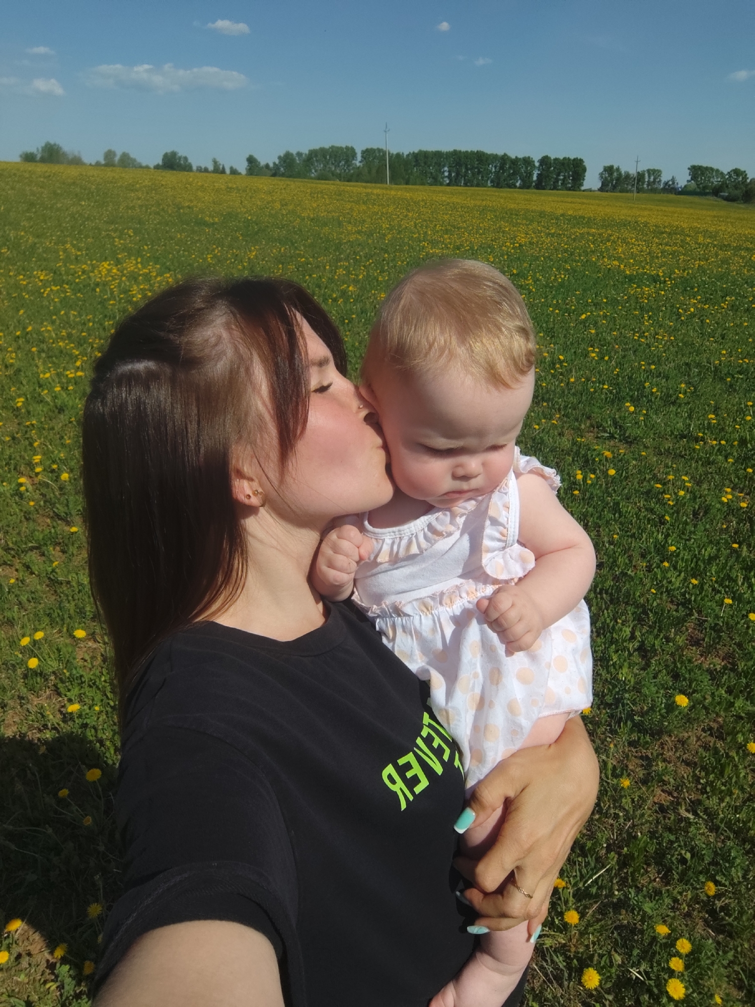 The image depicts a young woman holding a baby outdoors. The woman has brown hair and is wearing a black t-shirt with neon green text on it. She is kissing the baby on the cheek. The baby has light blonde hair and is wearing a white dress with orange polka dots. They are standing in a field of dandelions under a clear blue sky with some clouds. There's a line of trees along the horizon in the background. This location is not known to be in the city of Фоки, Russia.