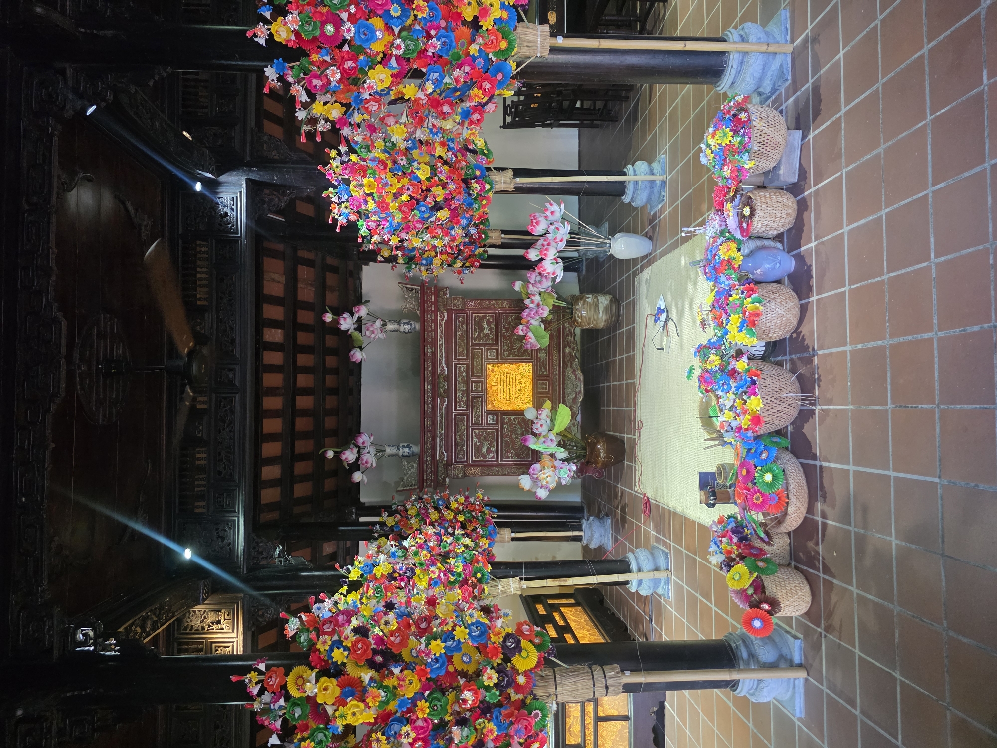 The image shows the interior of a building, possibly a temple or historical structure, in Đà Nẵng, Vietnam, characterized by a traditional architectural style with dark wooden beams and a red tiled floor. Vivid displays of colorful artificial flowers are prominent. The flower displays are in two arrangements, one overhead and another set of flower baskets at floor level. In the background is a central shrine or altar, adorned with gold accents, indicating a place of reverence. The lighting is a mix of natural and artificial. The perspective is from a relatively high vantage point, looking down into the space.