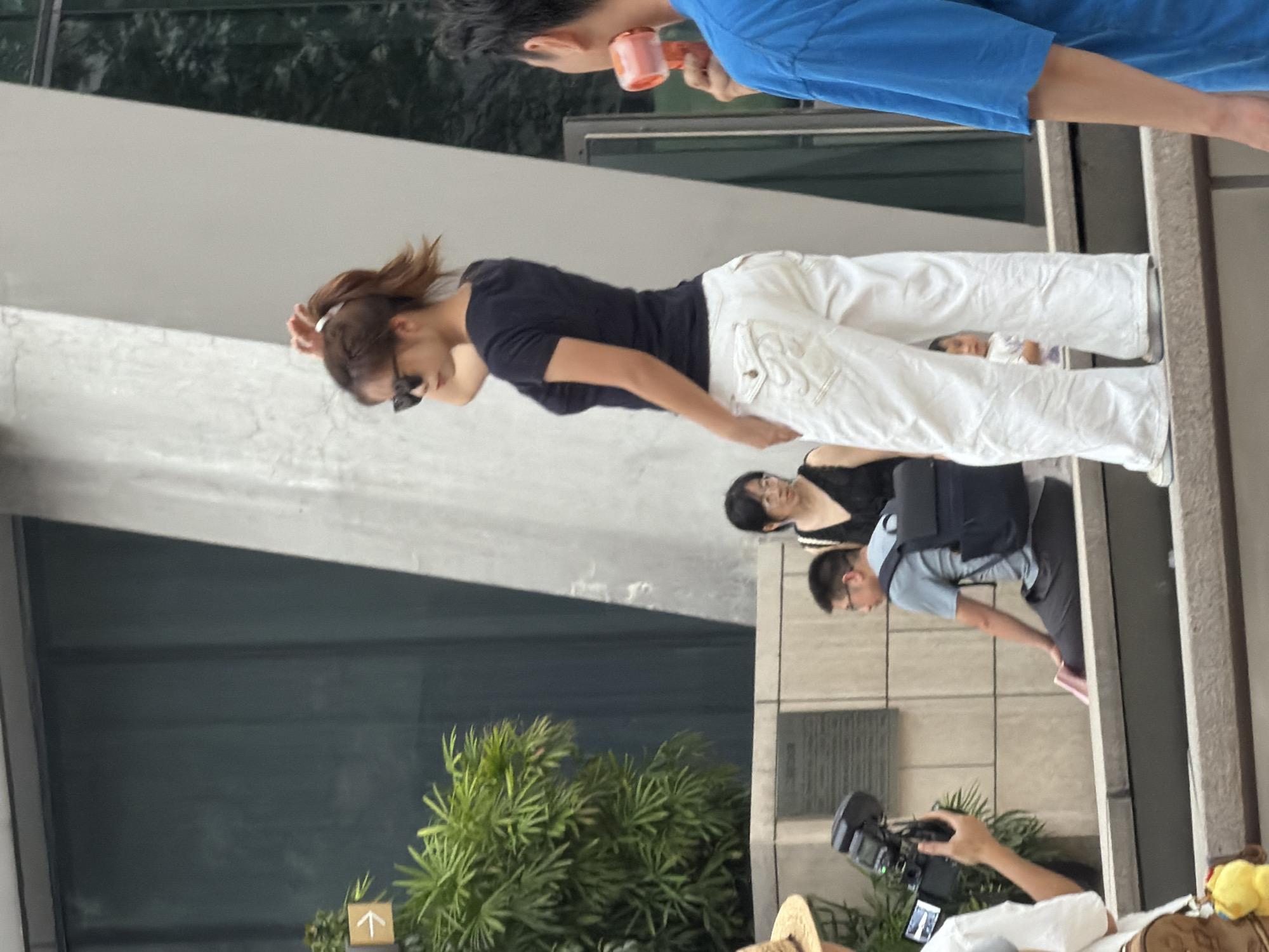 A woman wearing sunglasses, a dark blue t-shirt, and loose-fitting white pants stands with her right hand in her hair, facing right. She is positioned in the foreground, slightly off-center to the right. Behind her, a person in a blue t-shirt holds a pink object. To the left, a man in a grey t-shirt and dark pants with a blue backpack is seated, looking to his left. Another person, wearing glasses and a black dress, is behind the seated man. In the far left of the frame, a person with a straw hat is partially visible, holding a camera. The background consists of a textured concrete structure and a dark paneled wall. A green plant is visible to the left of the seated man. The setting appears to be an outdoor urban environment in Yuzhong District, China.