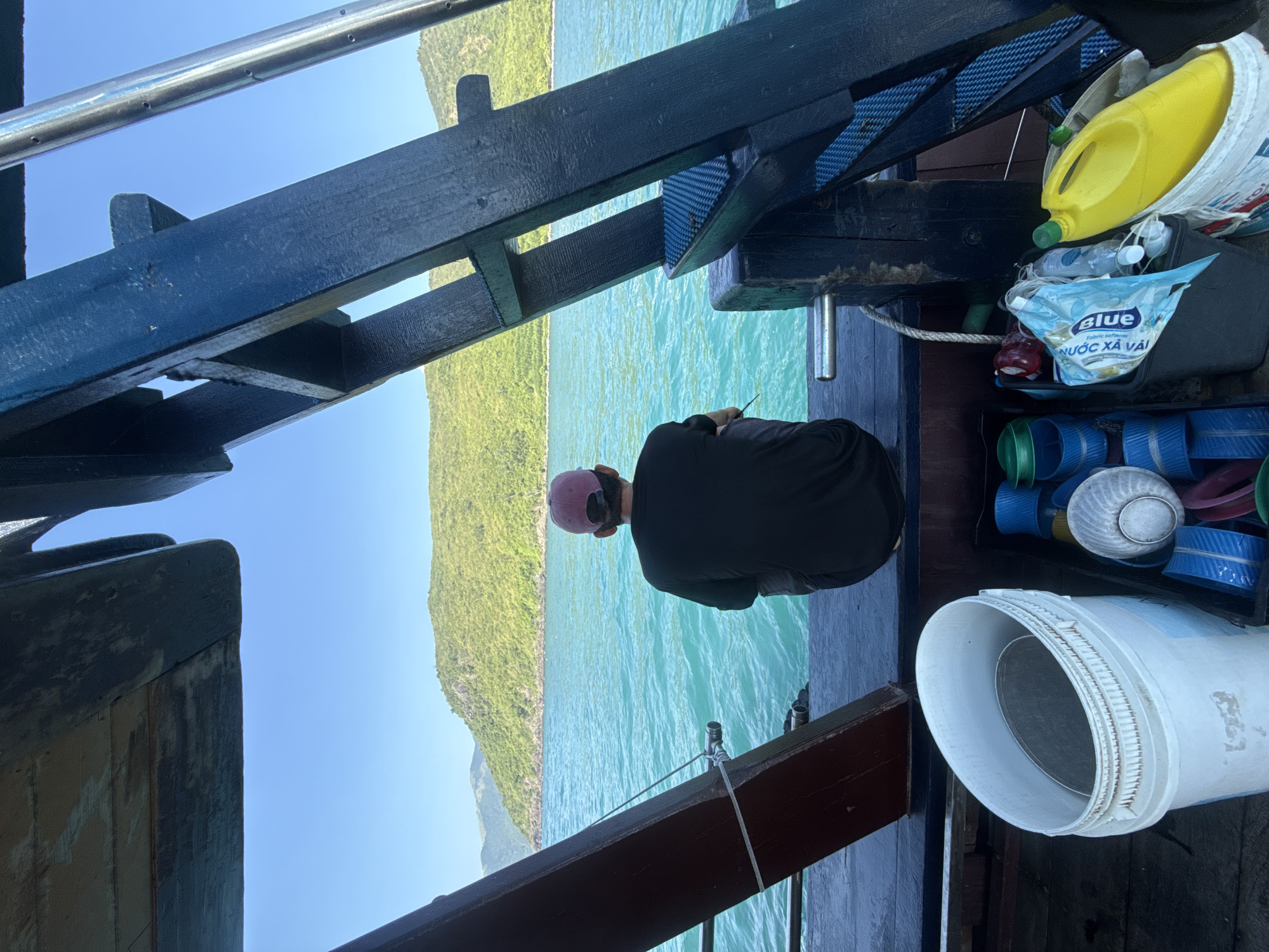 A person wearing a dark t-shirt and a pink cap is seated on the edge of a wooden boat, facing away from the camera. The person appears to be holding a fishing rod, casting into the turquoise water. The boat's wooden structure, painted a dark blue and brown, is visible in the foreground. Various containers, including a white bucket with a metal sieve inside and a yellow plastic jug, are stored on the boat's deck. In the background, a lush green island with a mountainous terrain rises from the calm sea under a clear blue sky. The scene suggests a tranquil fishing trip in the waters off Nam Nha Trang Ward, Vietnam.