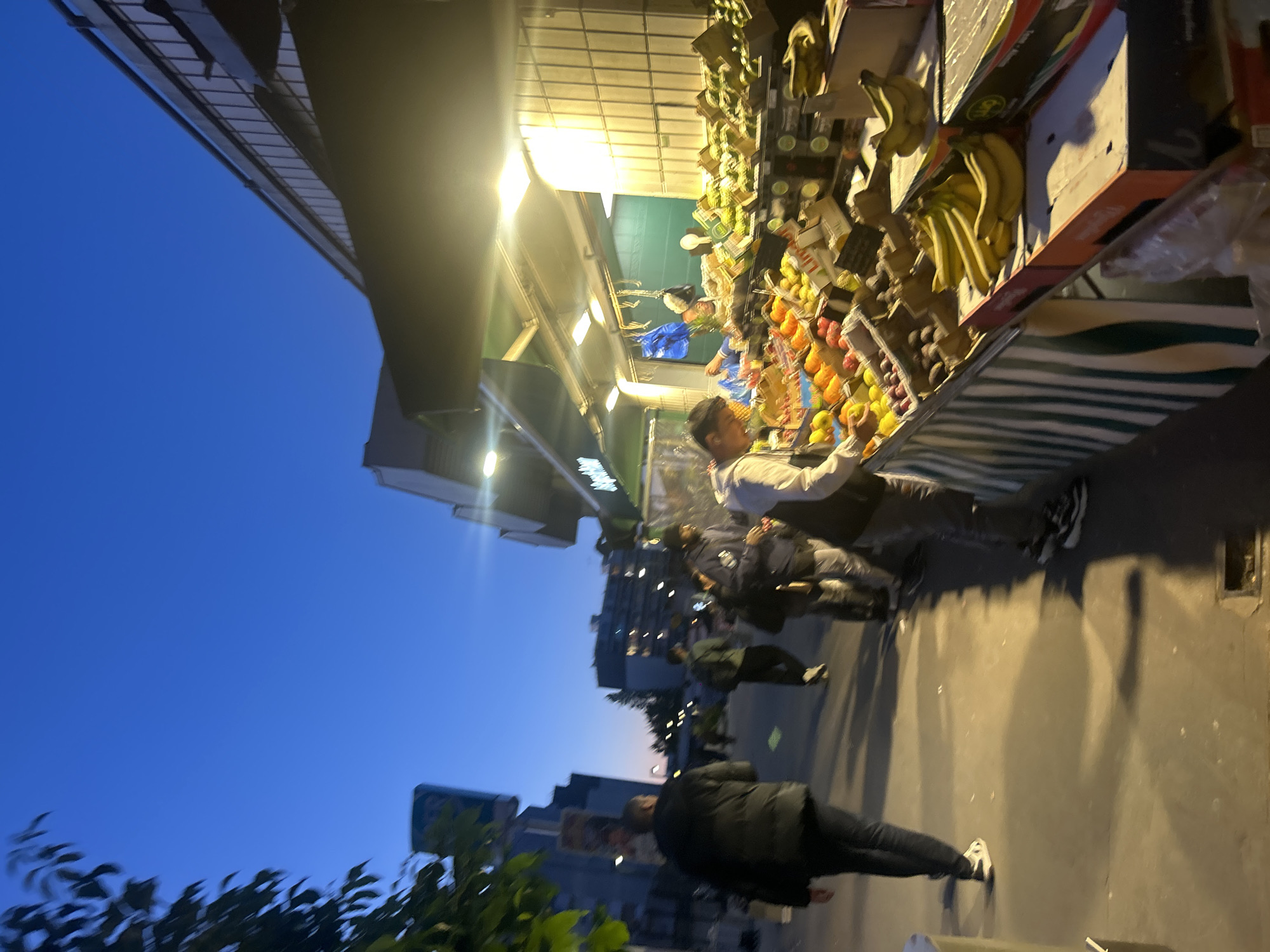 The image shows a street scene in Noisy-le-Grand, France, at dusk. A fruit stand is positioned on the right side, stocked with various fruits including bananas, oranges, lemons and avocados. A man is working at the stand, arranging or serving customers. Several pedestrians are walking along the sidewalk in front of the fruit stand. Buildings and trees are visible in the background against the blue sky.