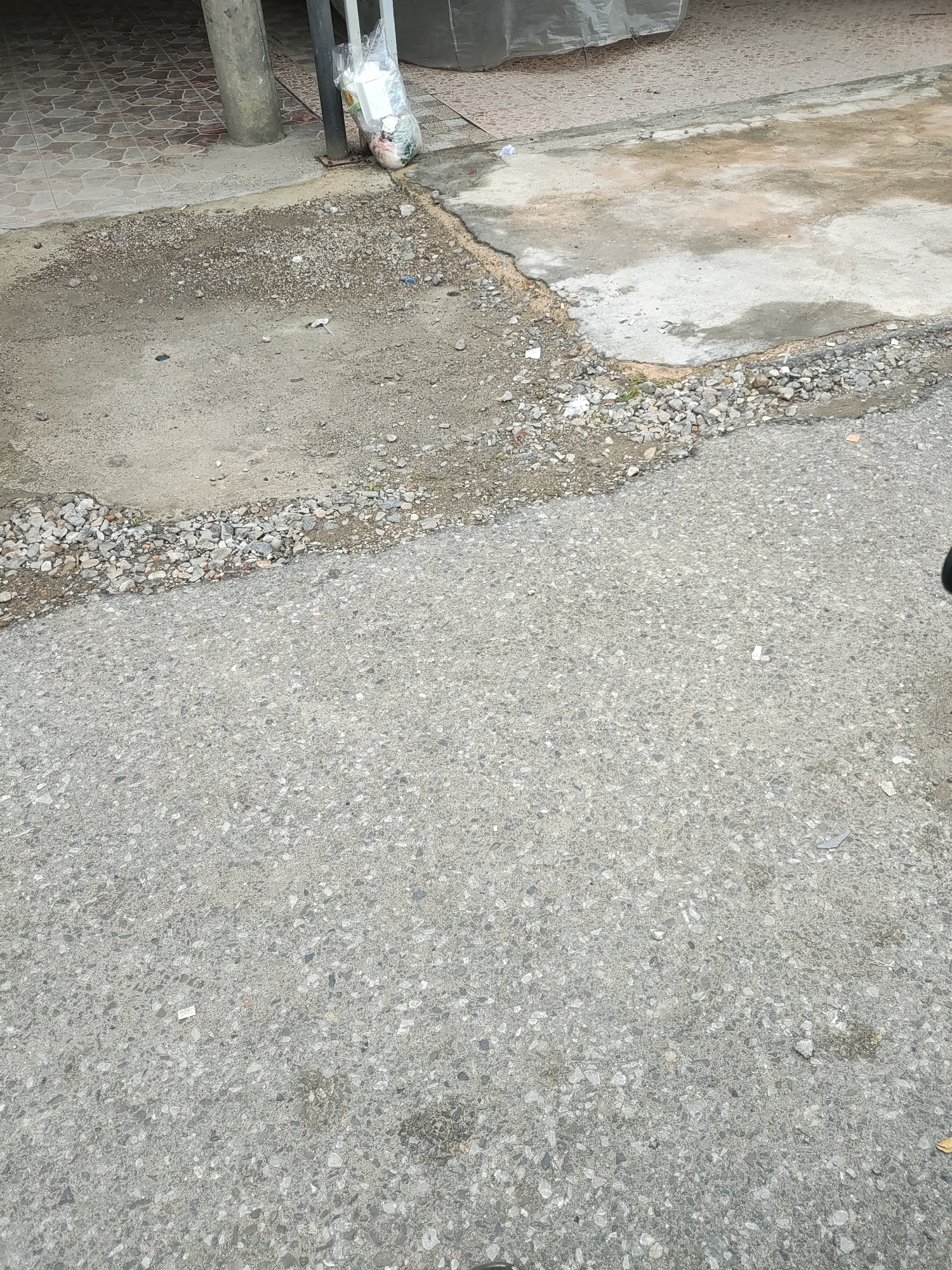 The image shows a portion of a street or road in Lhokseumawe, Indonesia. The surface is asphalt with a medium-grey tone. The foreground is mostly the street surface. The upper portion of the image shows a section of sidewalk or adjacent pavement, exhibiting a mix of textures and materials. A covered area, possibly a building overhang, can be seen at the top left. There is a trash bag located near a pillar. The transition between the road surface and the sidewalk is irregular, with small rocks and debris along the edge. No people, animals, or vehicles are visible in the frame. The lighting appears to be natural and overcast.